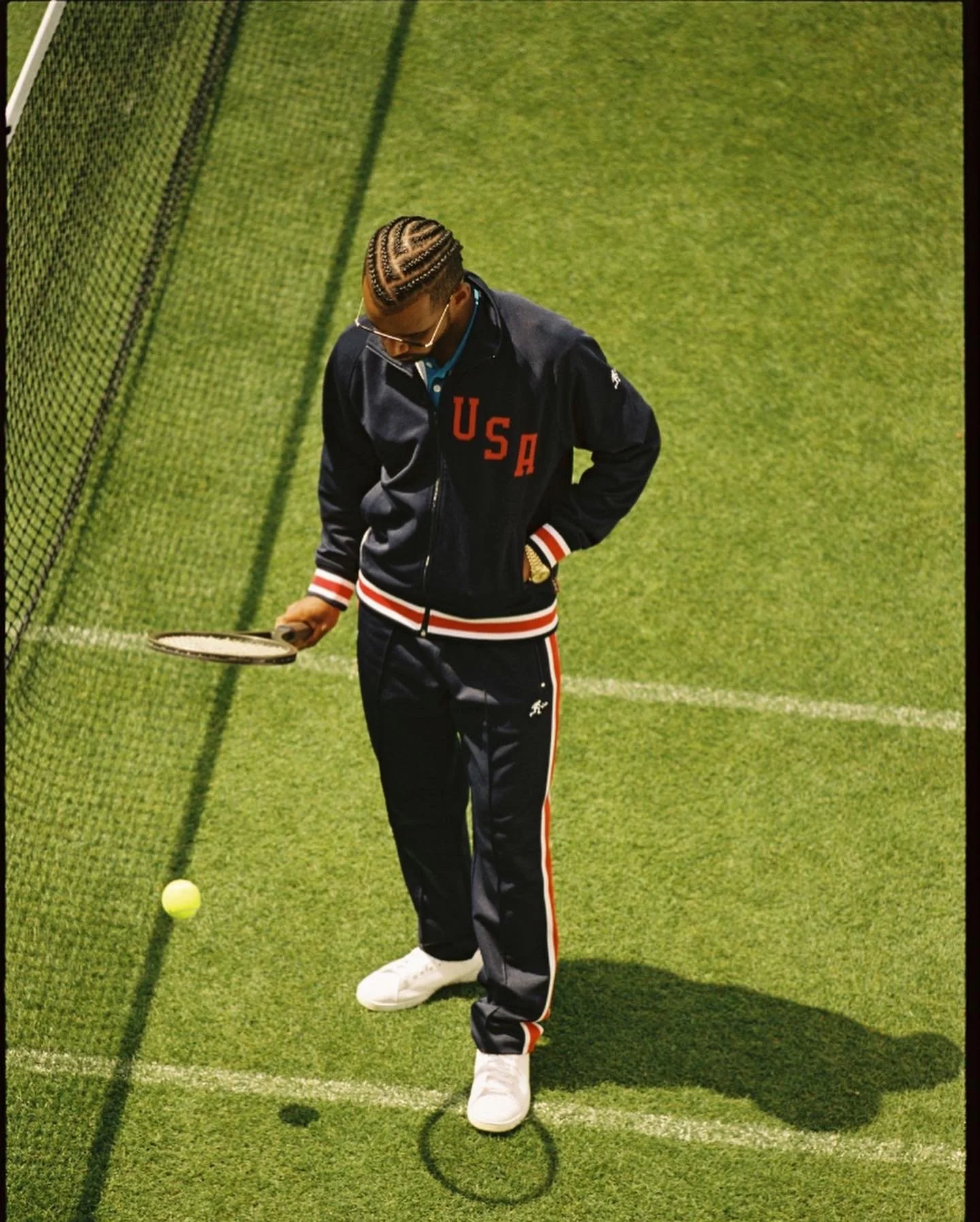 A man with braided hair, wearing sunglasses and a USA jacket, stands on a green tennis court holding a racket, looking down at a tennis ball.