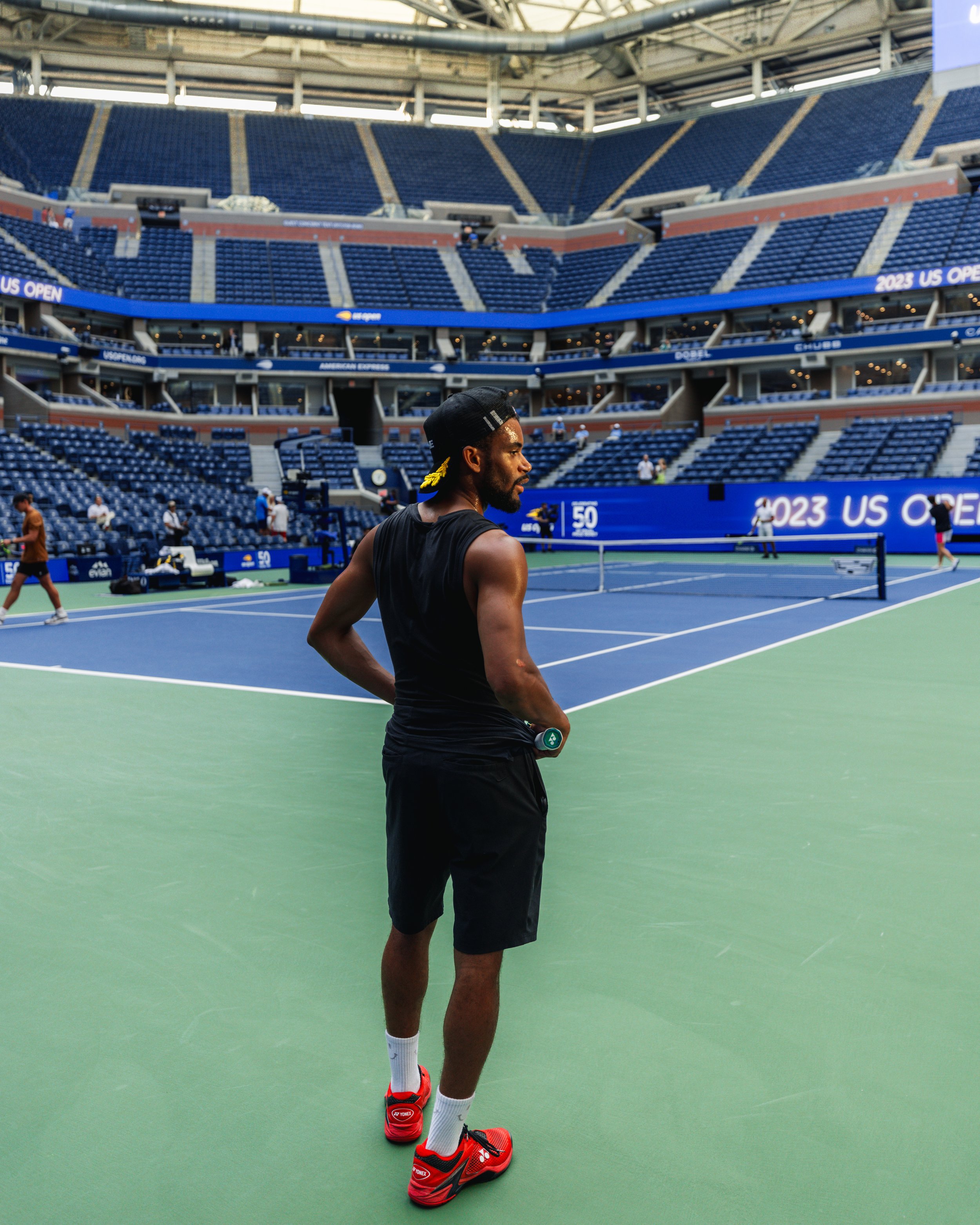 A professional male tennis player stands on a tennis court inside a large stadium, preparing for a match during the 2023 US Open.