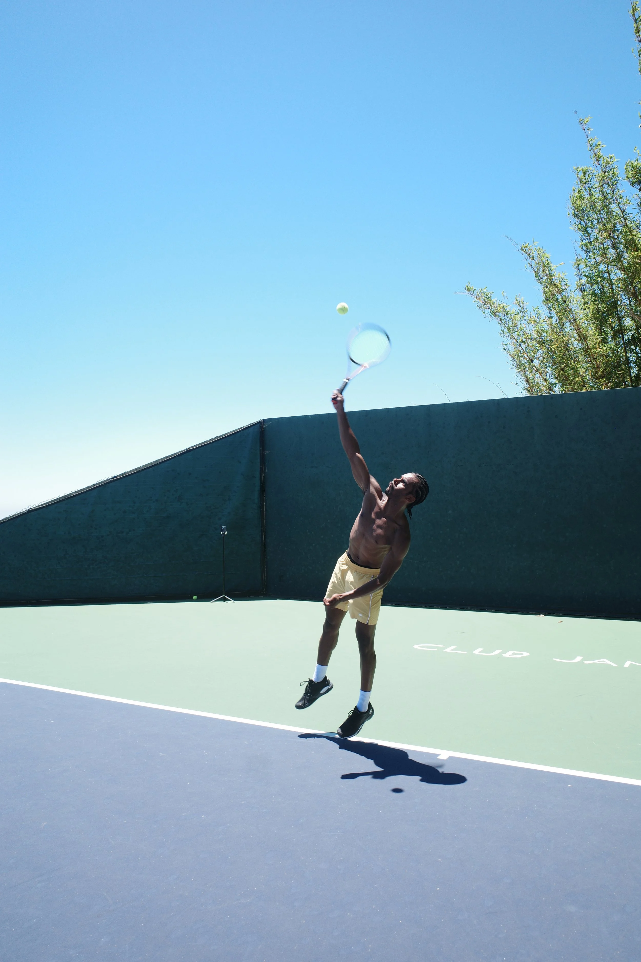 A shirtless man in yellow shorts and black sneakers jumping on a tennis court, hitting a tennis ball with a racket, with a clear blue sky and green trees in the background.