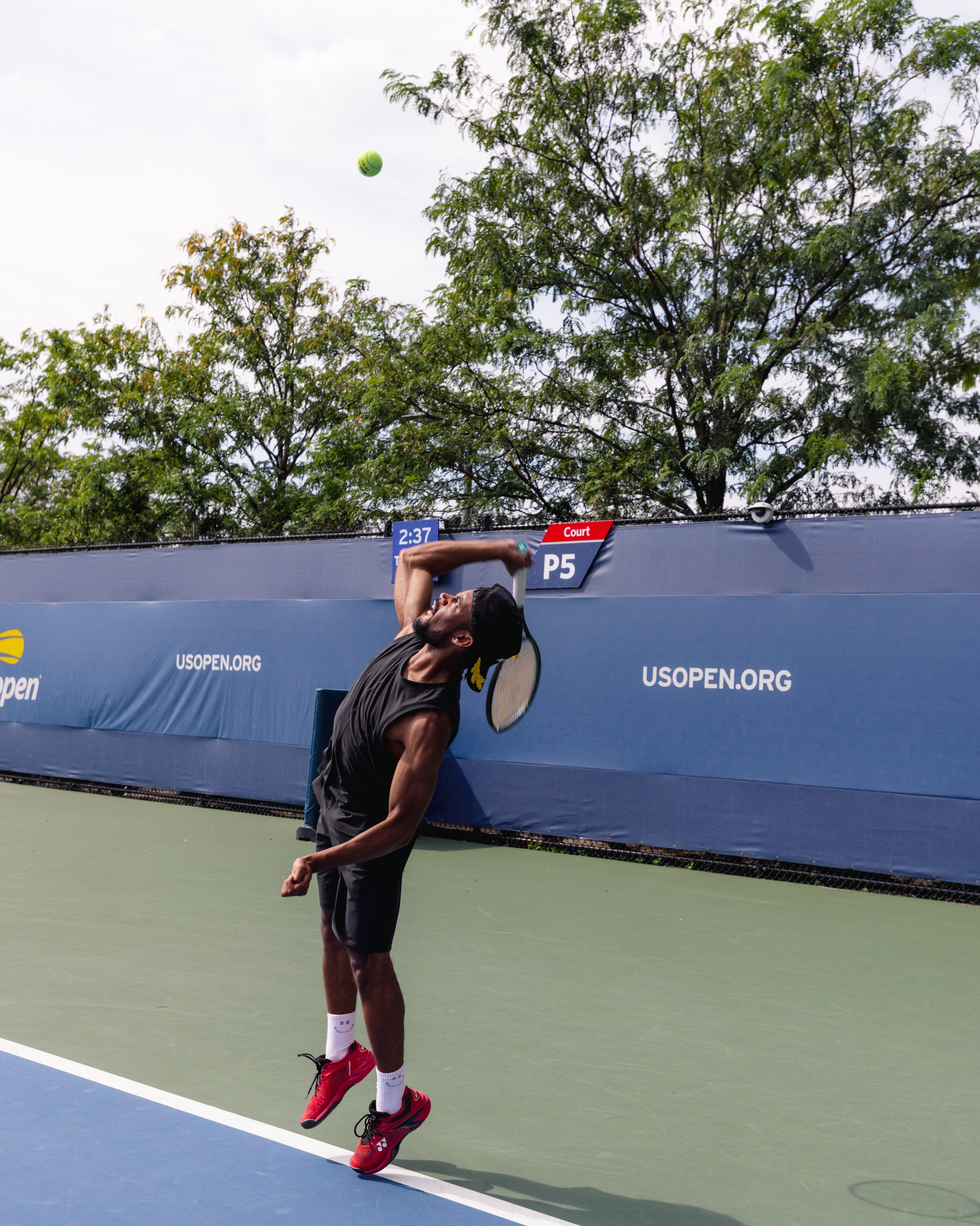 A man playing tennis on an outdoor court, about to hit a tennis ball, with trees in the background and a blue US Open banner along the fence.