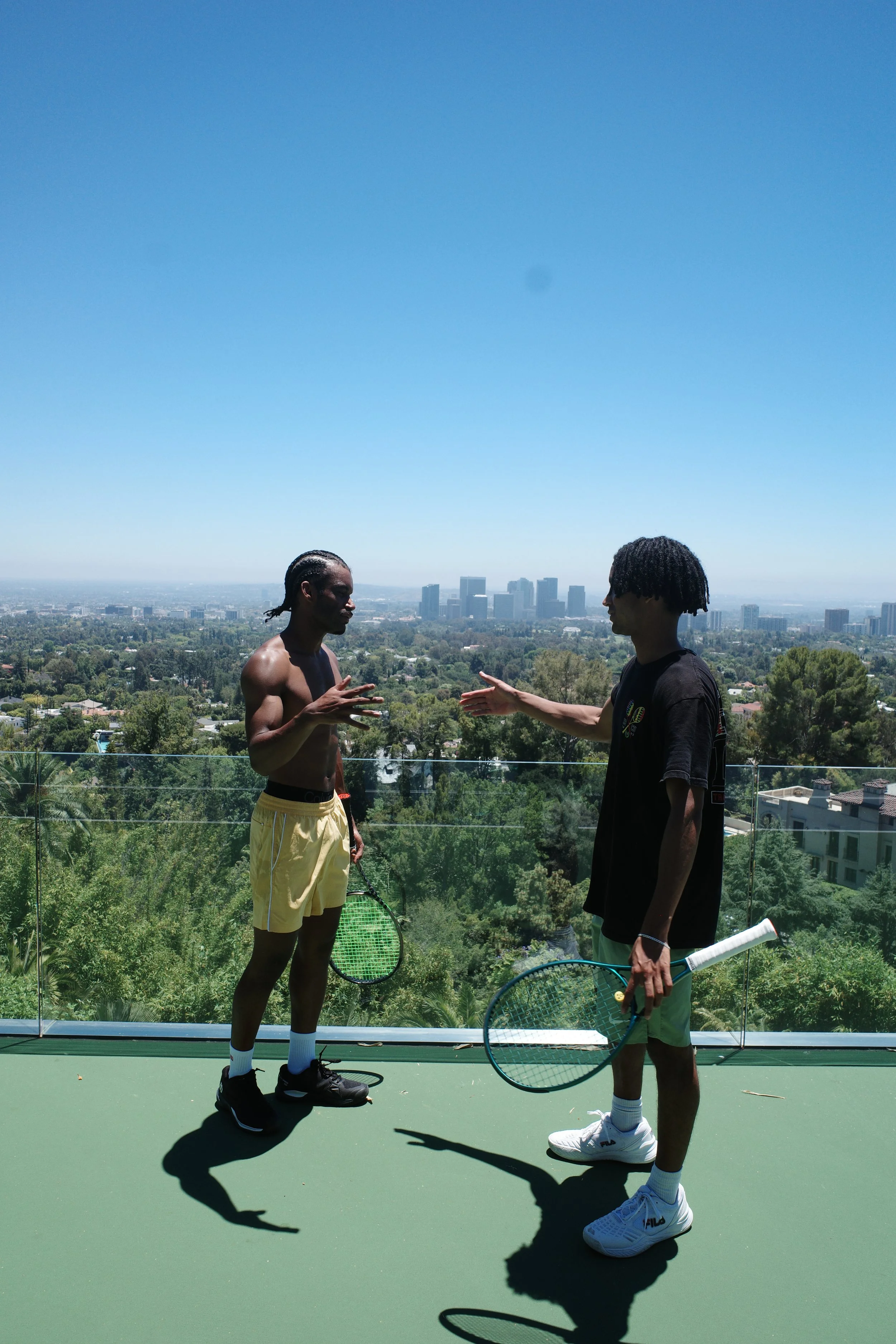 Two young men holding tennis rackets on a rooftop tennis court with a city skyline and trees in the background.