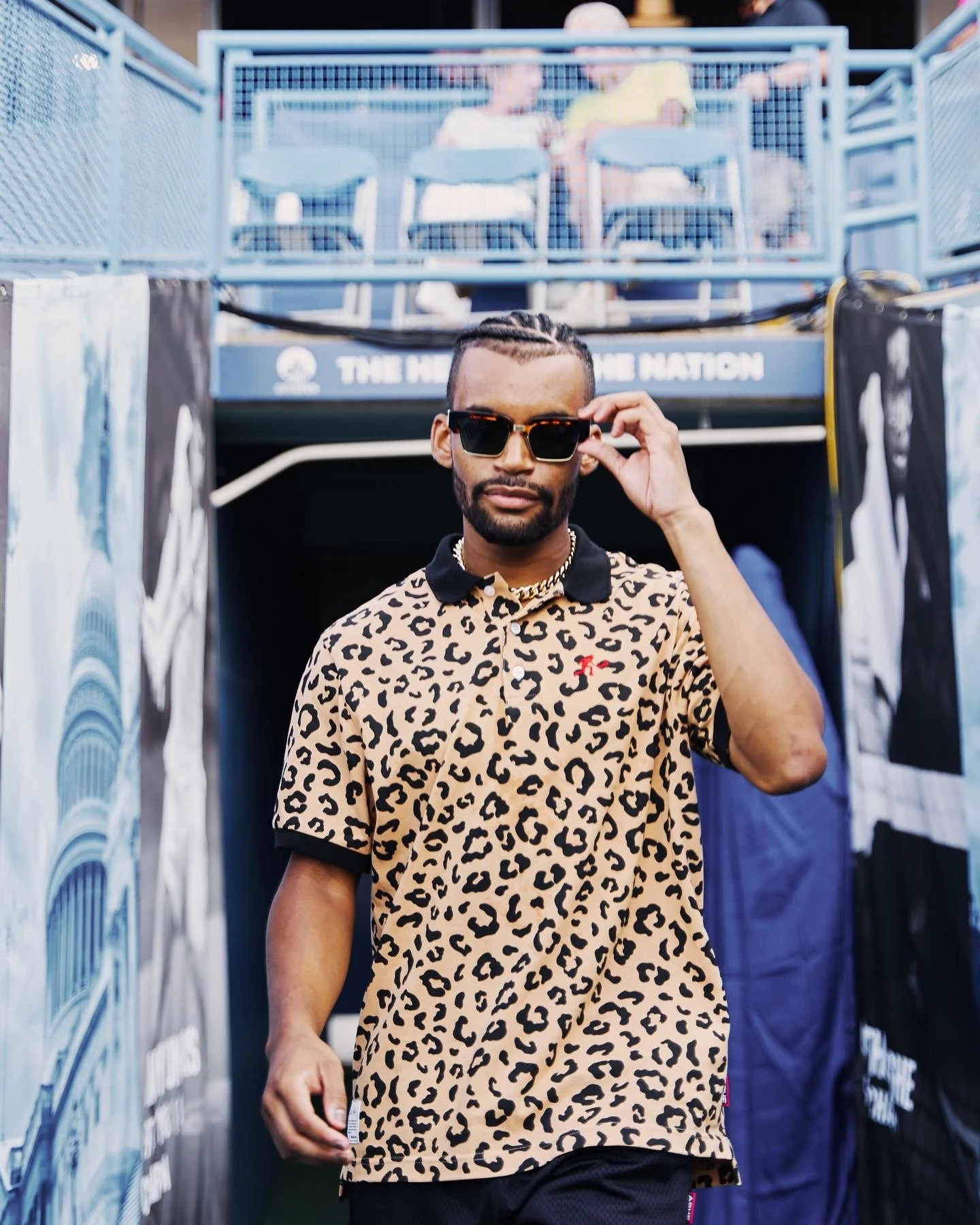 Man wearing sunglasses and a leopard print polo shirt walking past a sports stadium entrance with banners.
