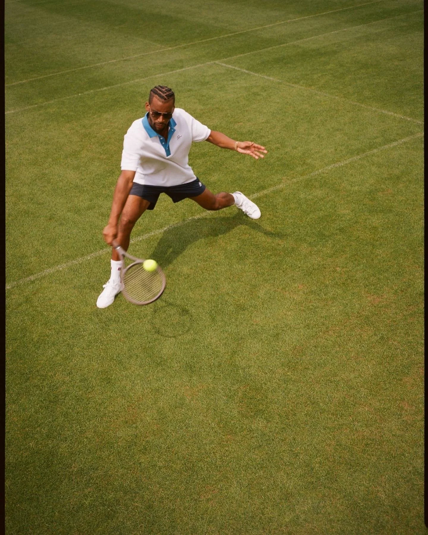 A man on a tennis court, wearing sunglasses, a white polo shirt, black shorts, and white sneakers, is hitting a tennis ball with a tennis racket.