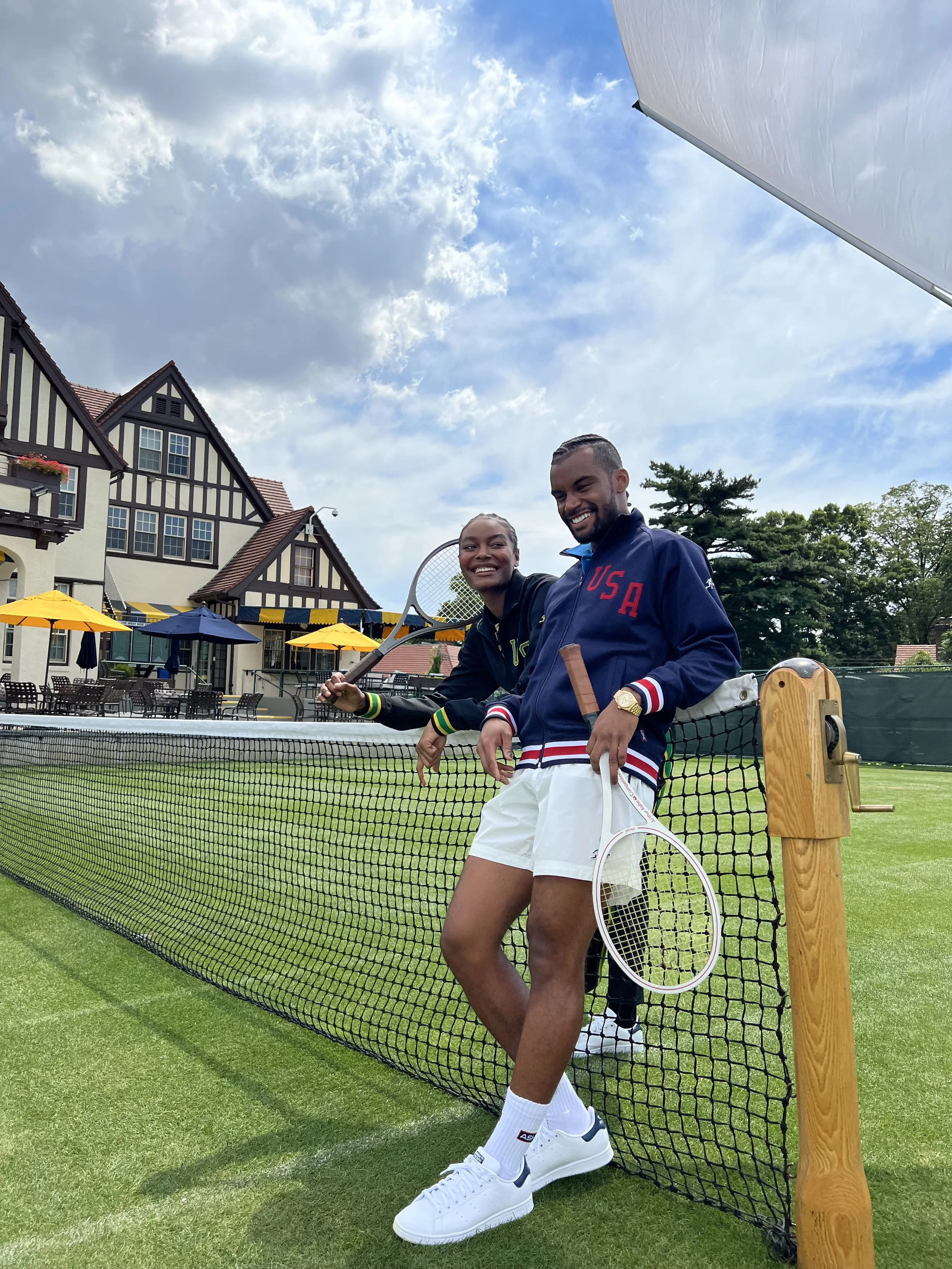 Two young adult tennis players, a female and a male, smiling and leaning on a tennis net on a grass court, with charming houses, outdoor seating with yellow umbrellas, and a partly cloudy sky in the background.