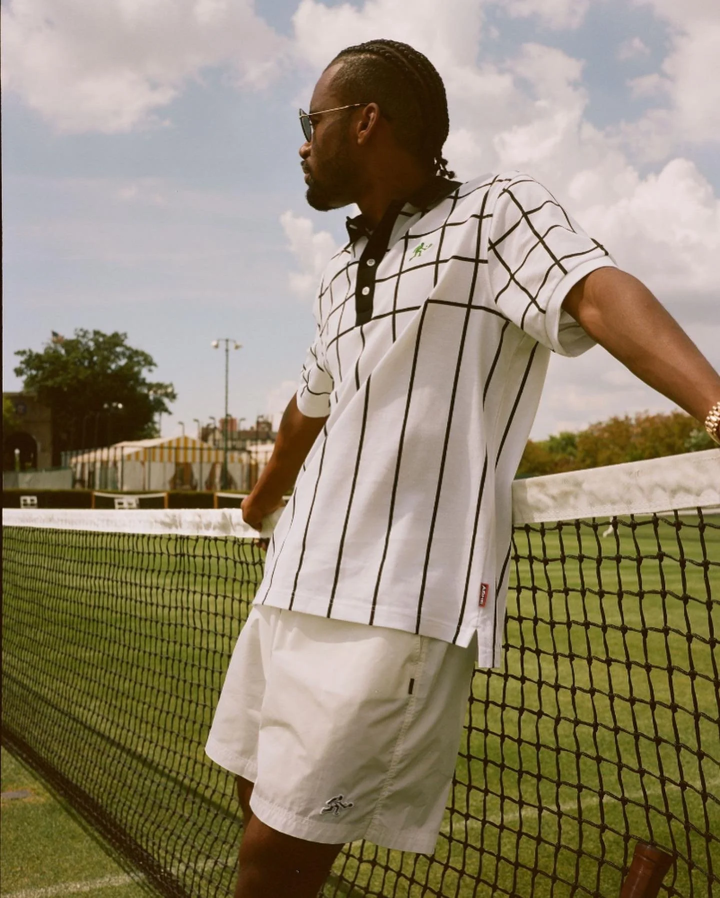 A man wearing sunglasses and a white polo shirt with a black grid pattern leans against a tennis net on a grassy tennis court during daytime, with trees and buildings in the background and a partly cloudy sky.