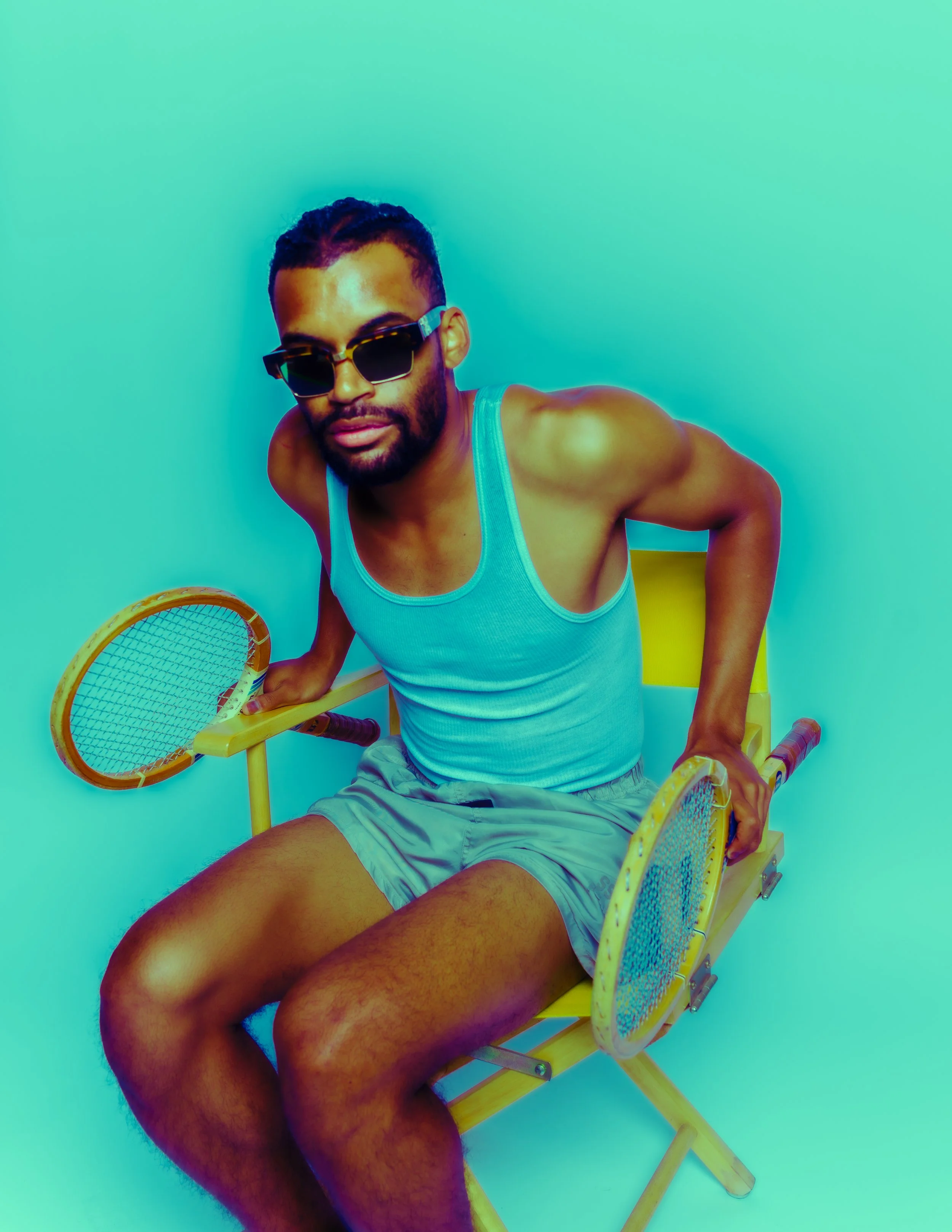 A man with dark hair, beard, and sunglasses sitting on a yellow chair against a turquoise background, holding tennis rackets in both hands.