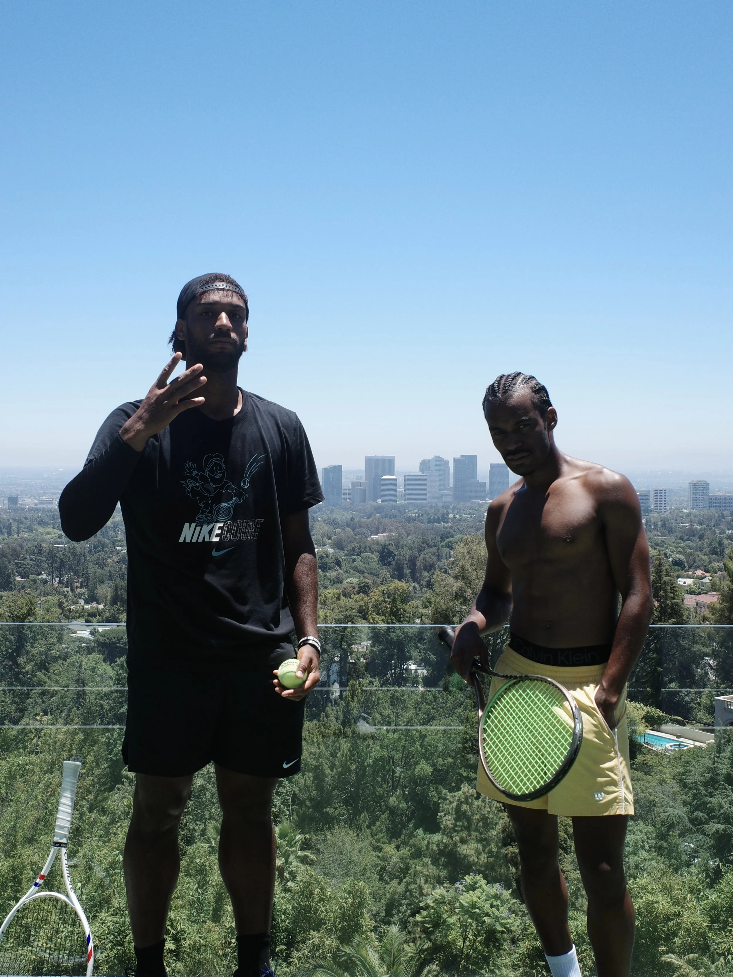Two young men standing outdoors on a sunny day, with a city skyline and green trees in the background. One is shirtless and holding a tennis racket, the other is wearing a black t-shirt and shorts, holding a tennis ball.