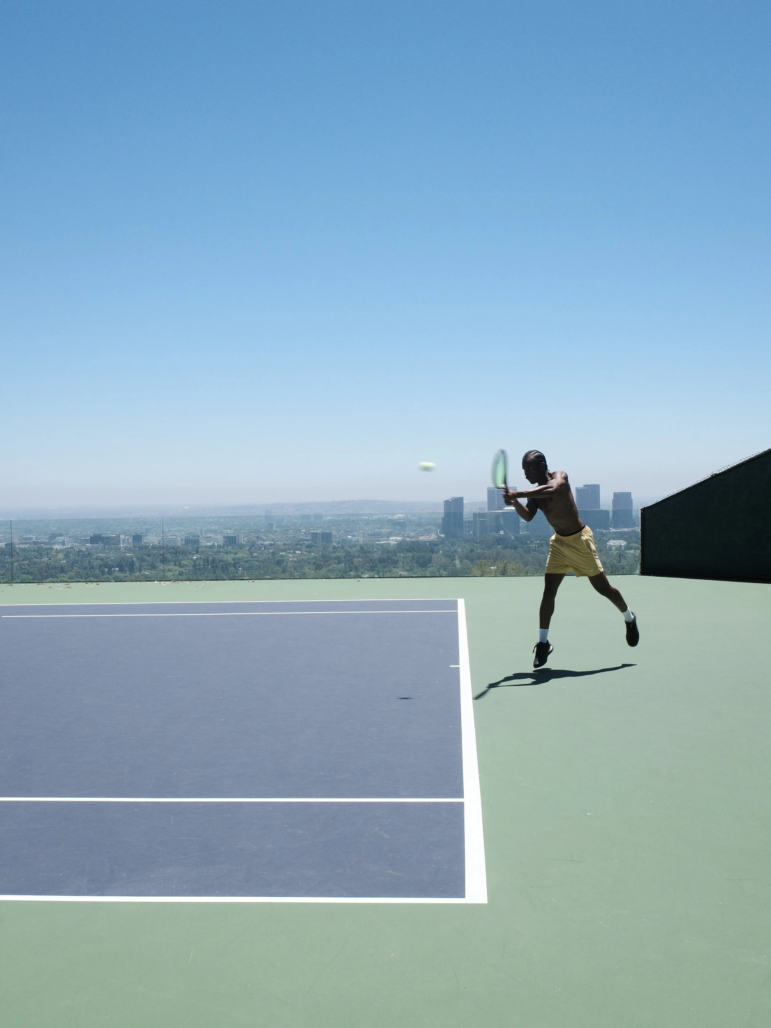 A person playing tennis on a rooftop court against a city skyline and blue sky.