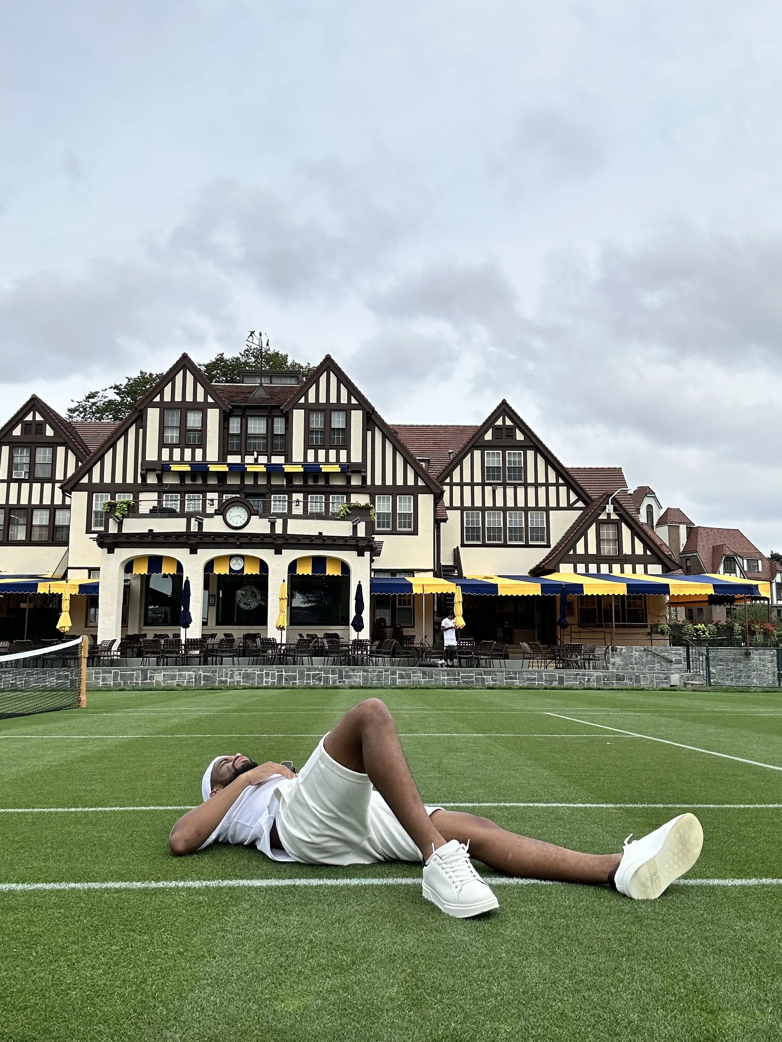 A man lying on a tennis court with a large house or resort in the background under a cloudy sky.