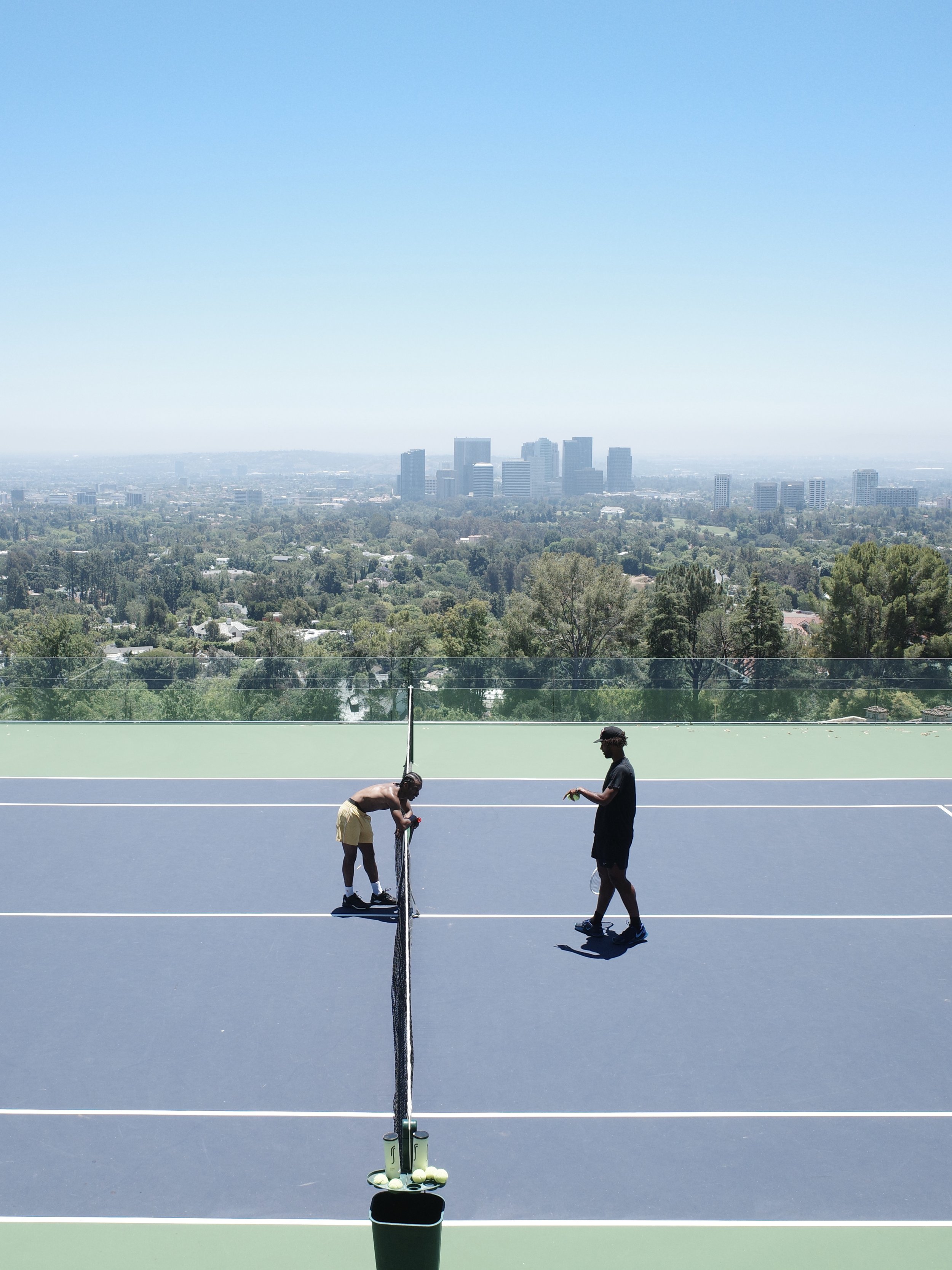 Two young men on a tennis court, one setting up or repairing the net, the other standing nearby holding a tennis racket, with a city skyline in the background.