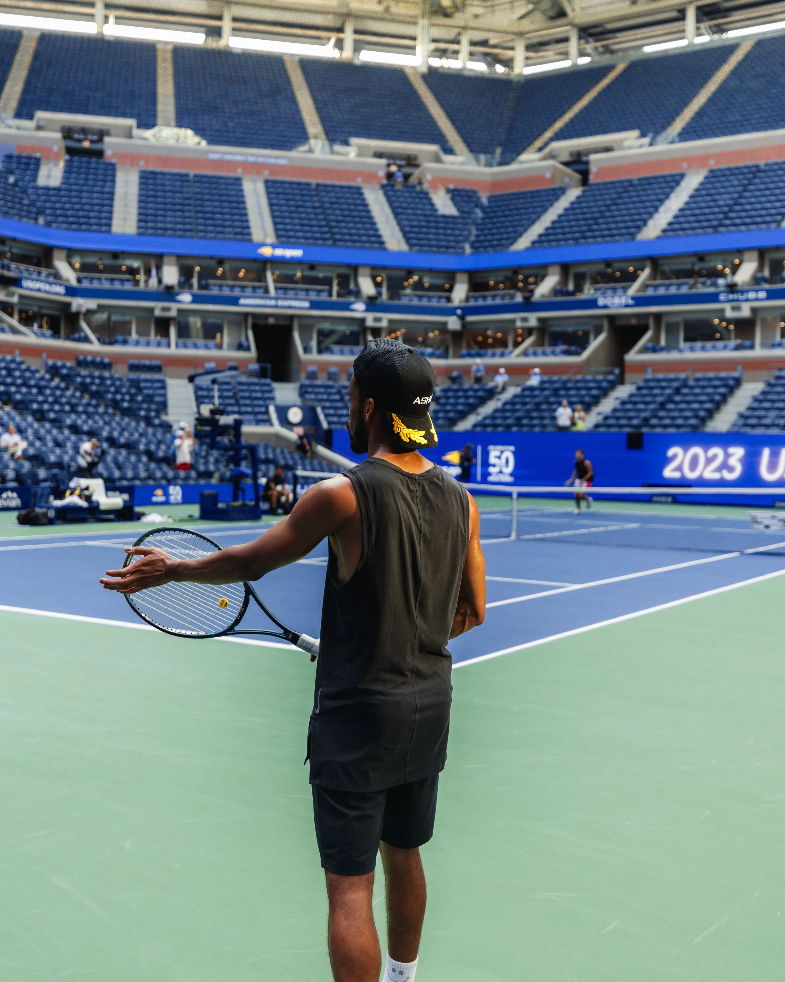 A tennis player stands on a tennis court holding a tennis racket, with an empty stadium surrounding him.