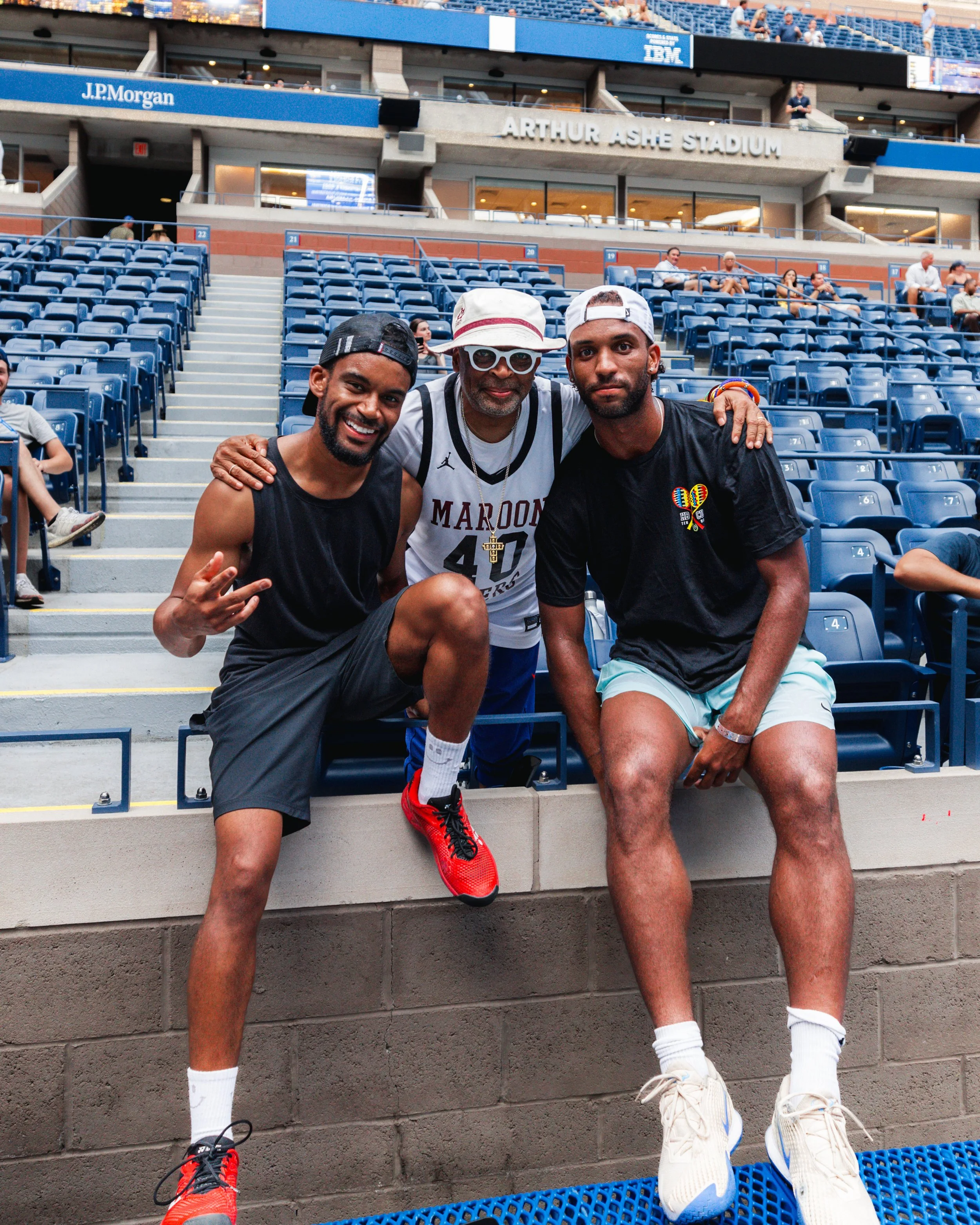 Three men posing together in a stadium, with empty blue seats behind them, including a sign that reads 'Arthur Ashe Stadium'.