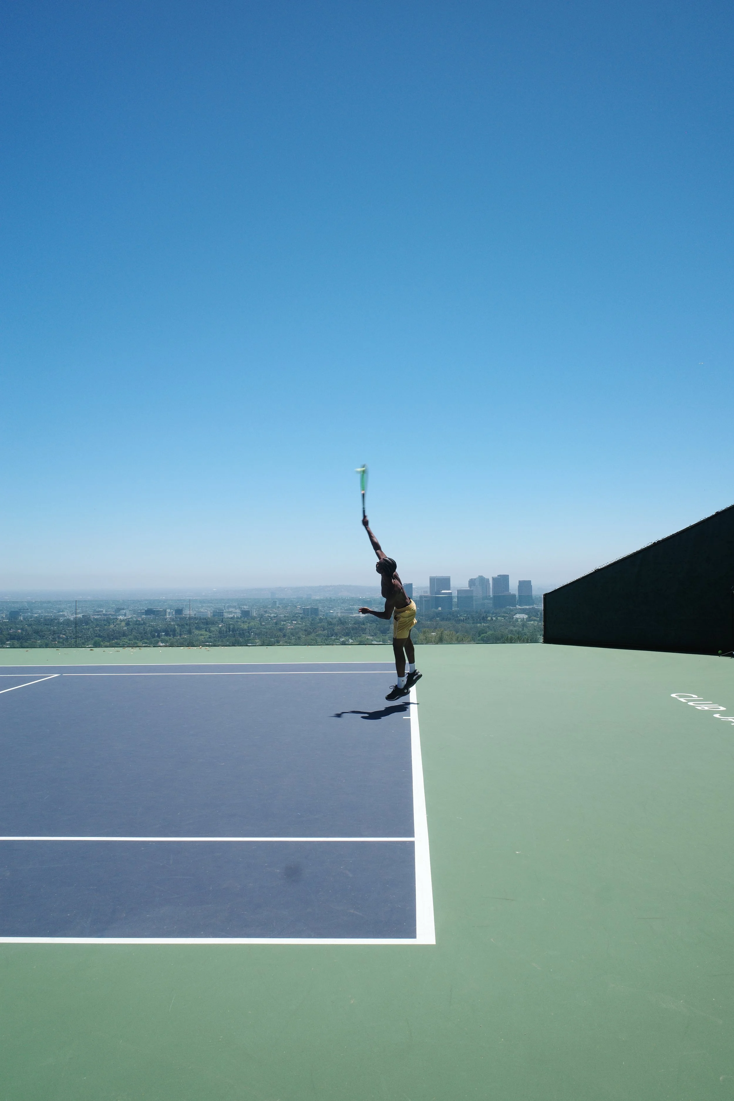 A person wearing yellow shorts and a black shirt playing tennis on a rooftop court with a city skyline in the background under a clear blue sky.