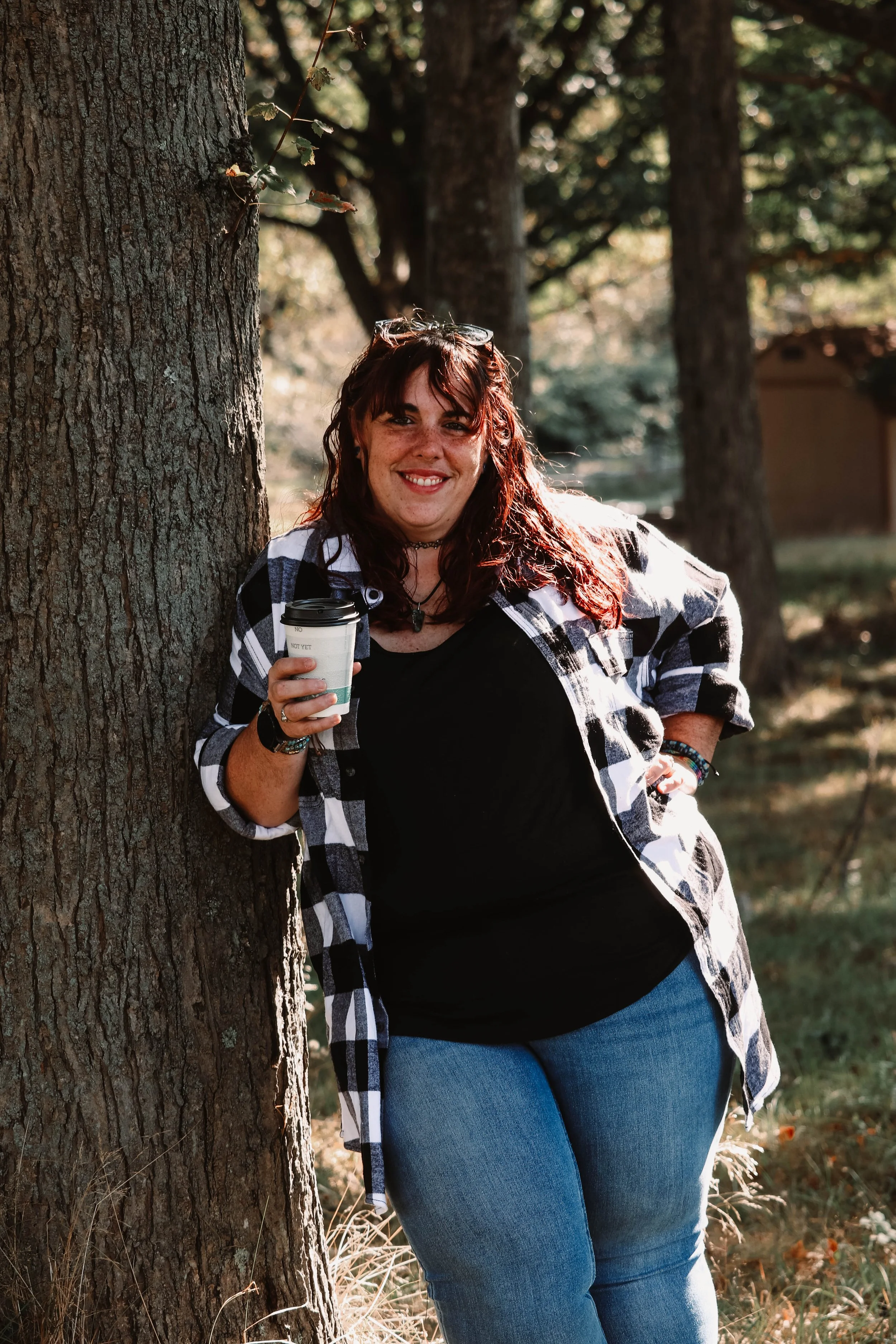 Kaylene holding a coffee, leaning against a tree.