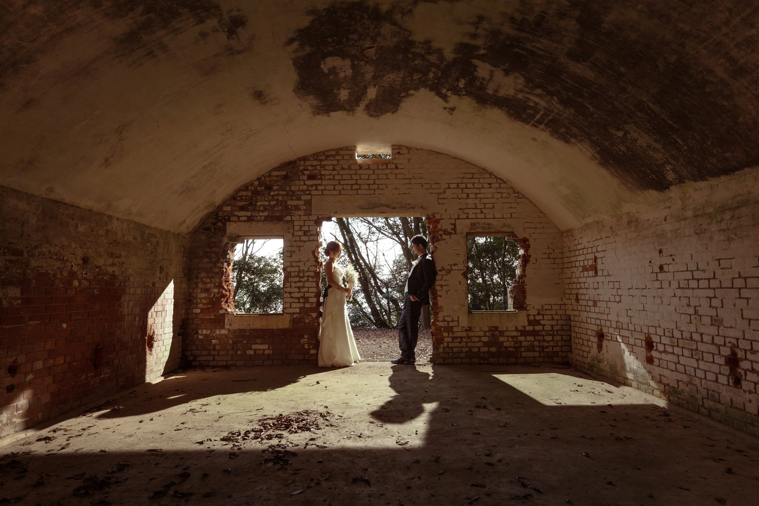 ロケ撮影イメージ | 江田島 三高山砲台跡 | 崩壊した壁のある古い建物の内部で、結婚式のカップルが向かい合って立っている。外の木々や空が見える背景が見える。