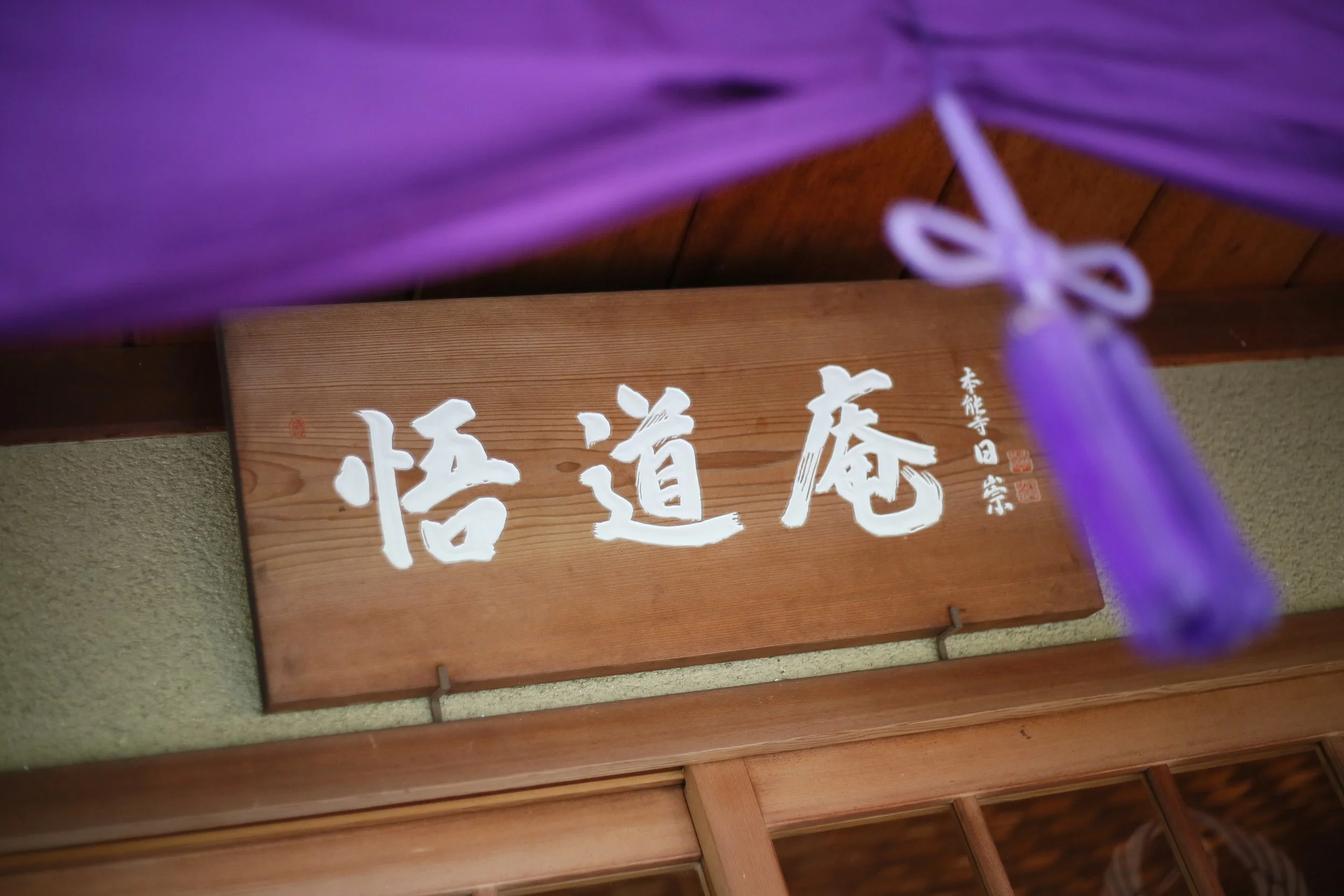 悟道庵点景 | 木製の看板に白い文字で書かれた「悟道庵」の文字。紫色の布の一部が上部に見える。