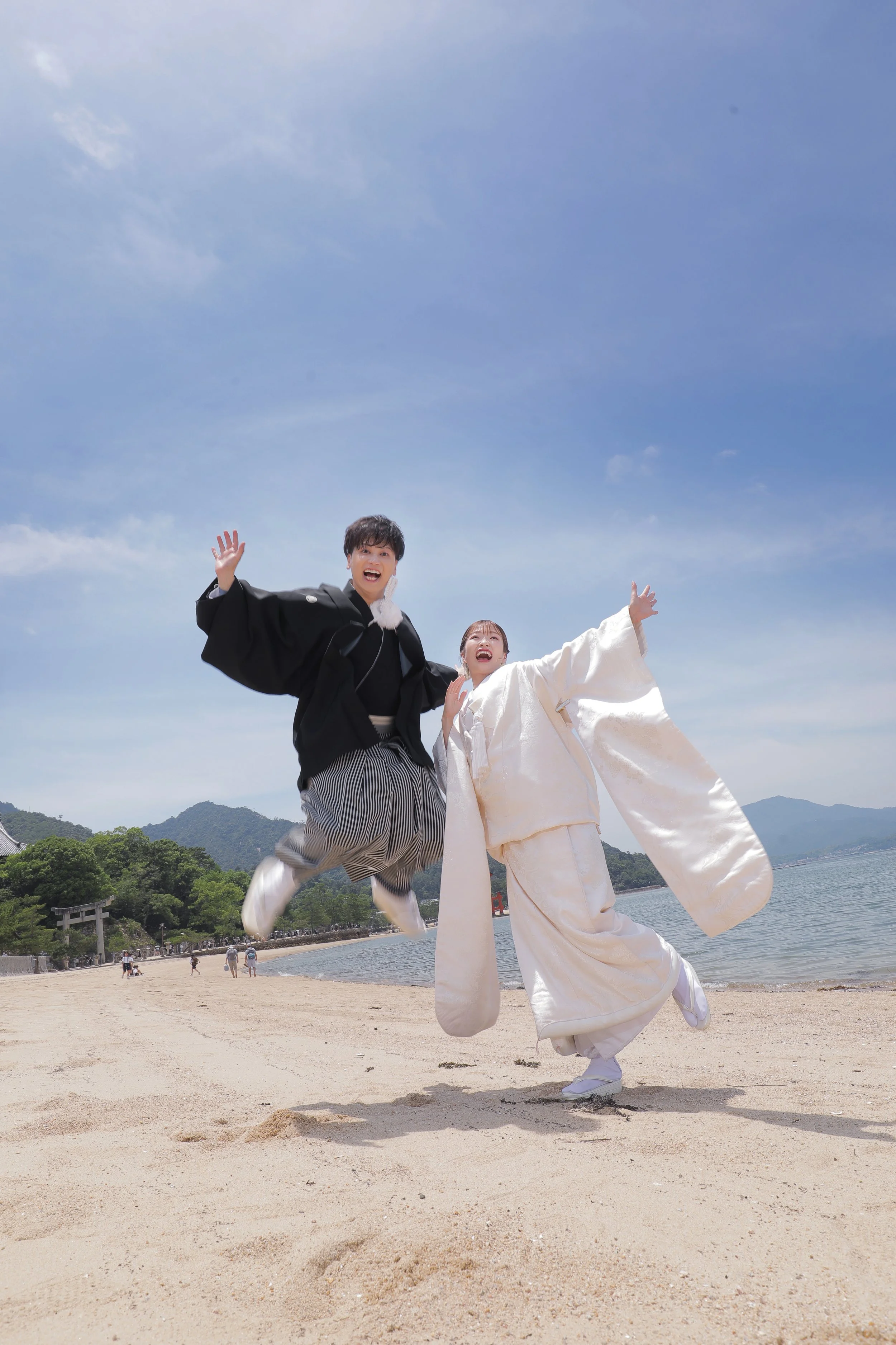宮島フォトギャラリー｜海辺でジャンプしている新郎新婦。青空と海が背景に広がり、遠景には大鳥居が見える。