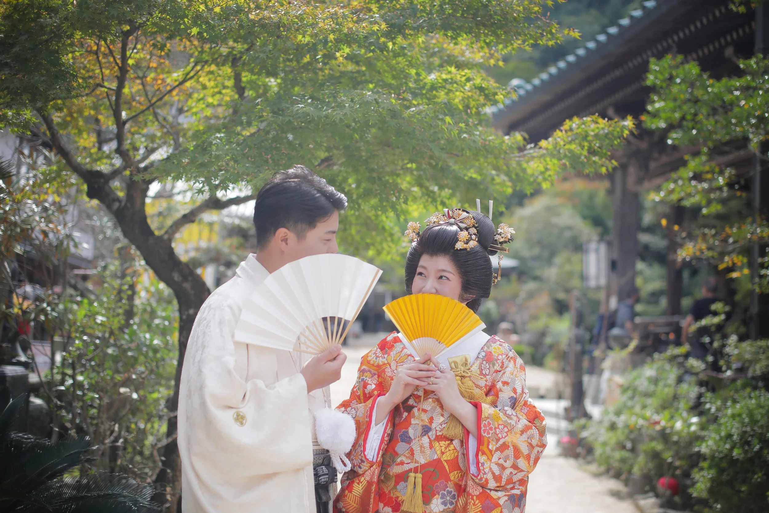 宮島フォトギャラリー｜大聖院ロケ｜新郎新婦が屋外でお互いに笑顔を交わしながら扇子を持っている光景