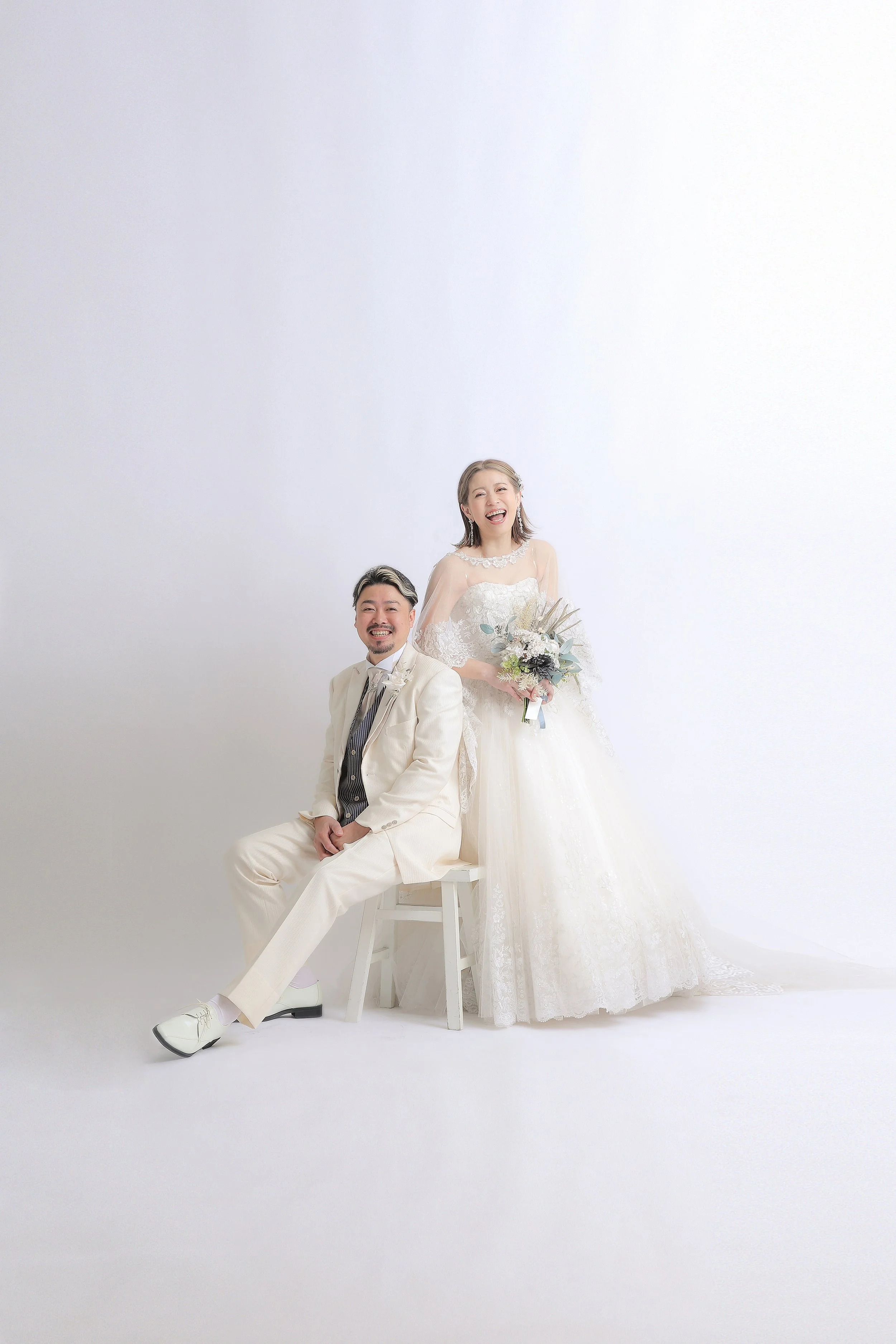 スタジオ撮影イメージ｜白い背景の前で笑顔の新郎と新婦の結婚式写真。新郎は白いスーツを着て座っており、新婦はホワイトのドレスを着て花束を持っている。
