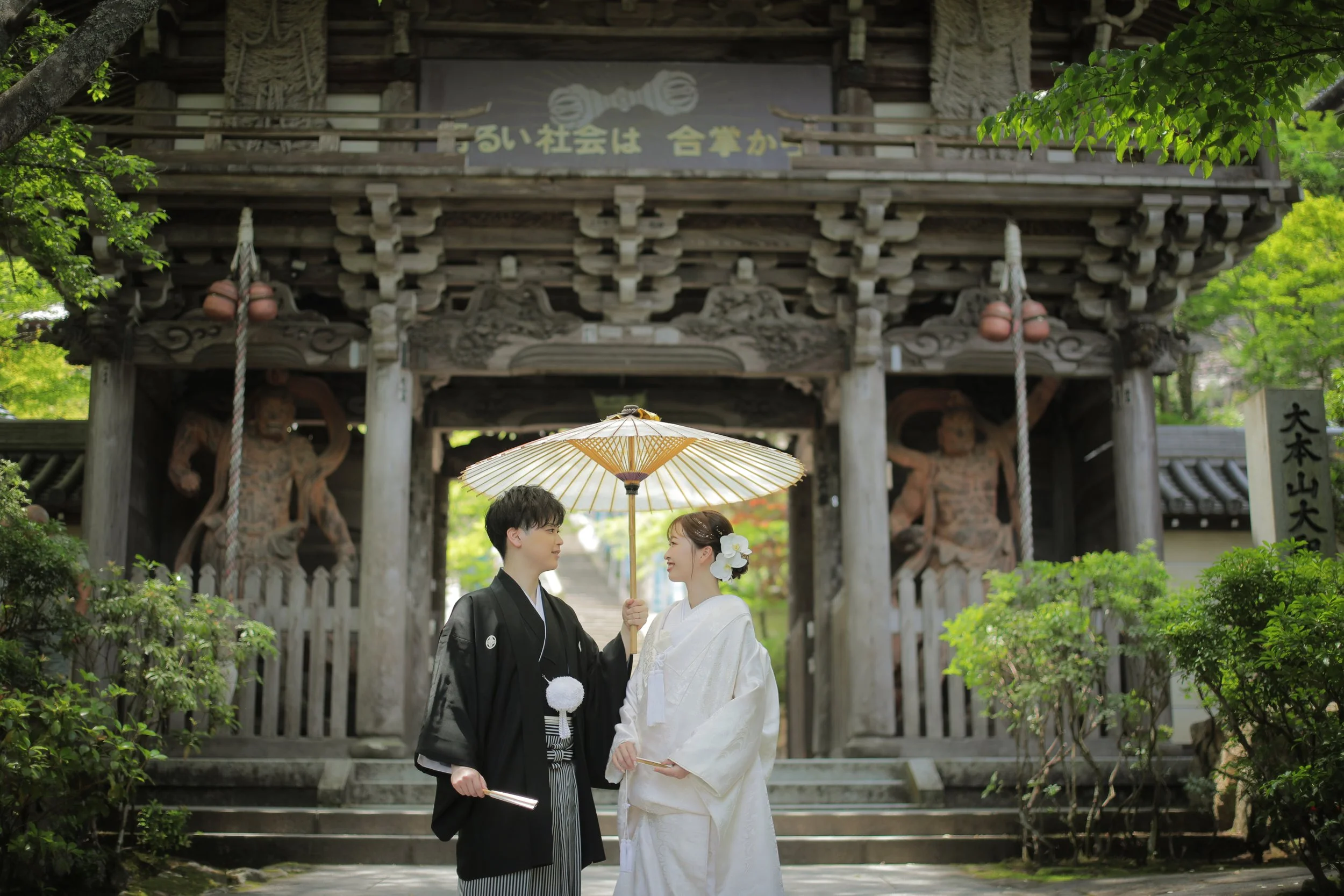 宮島フォトギャラリー｜大聖院ロケ｜大聖院山門前で傘を持ちながら微笑み合う新郎新婦。