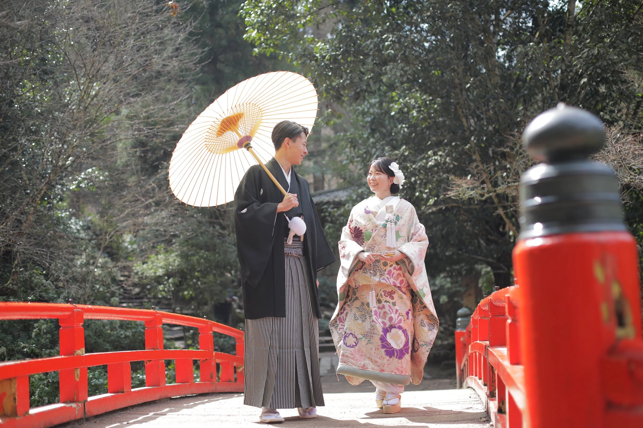 宮島フォトギャラリー｜もみじ橋を渡る新郎新婦。和傘を持ちながら笑顔で会話している。周囲は自然の景色が広がっている。