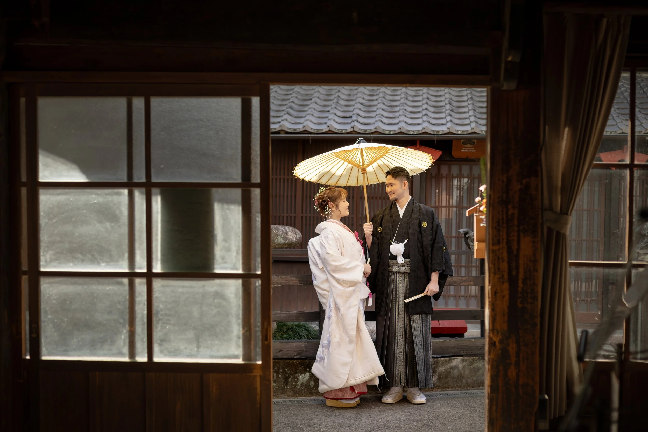 ロケ撮影イメージ｜御手洗町並み保存地区｜和装の新郎新婦が外で出会い、和傘を持って微笑み合っている場面。背景には木造の建物と屋根が見える。