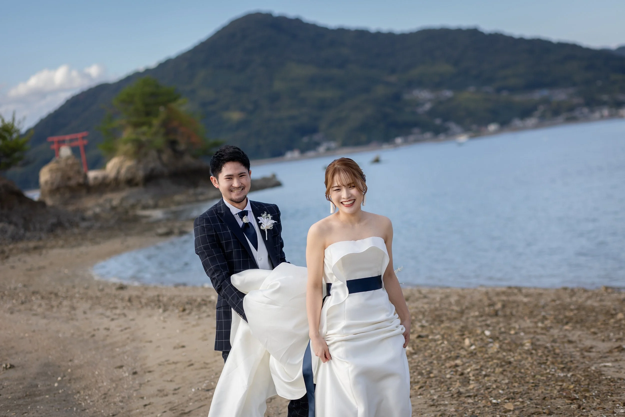 ロケ撮影イメージ | 江田島 ネイビーロード | 海辺の新郎新婦。新婦は白いドレスを着て笑顔、花嫁のドレスは黒い帯付き。新郎は黒いタキシード、笑顔で新婦に手を差し伸べている。背景には海と山と赤い鳥居が見える。