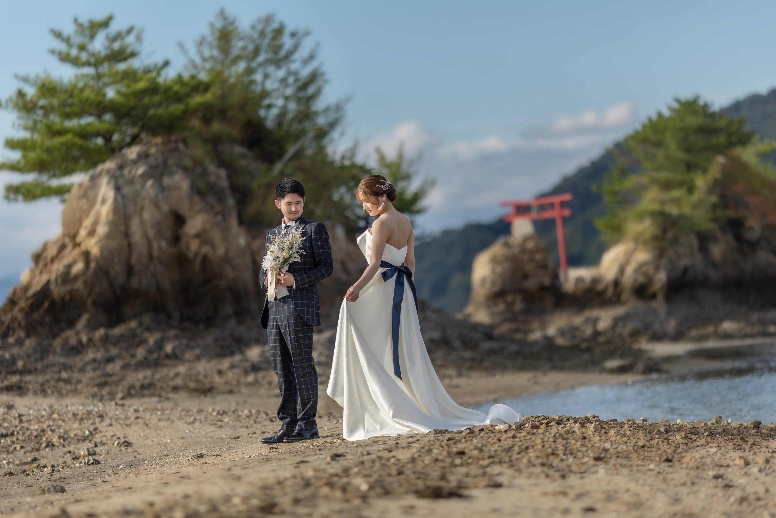ロケ撮影イメージ | 江田島 ネイビーロード | 海辺の新郎新婦。男性は紺色のチェック柄スーツ、女性は白のドレスを着用。背景に岩と木のある島と鳥居が見える。