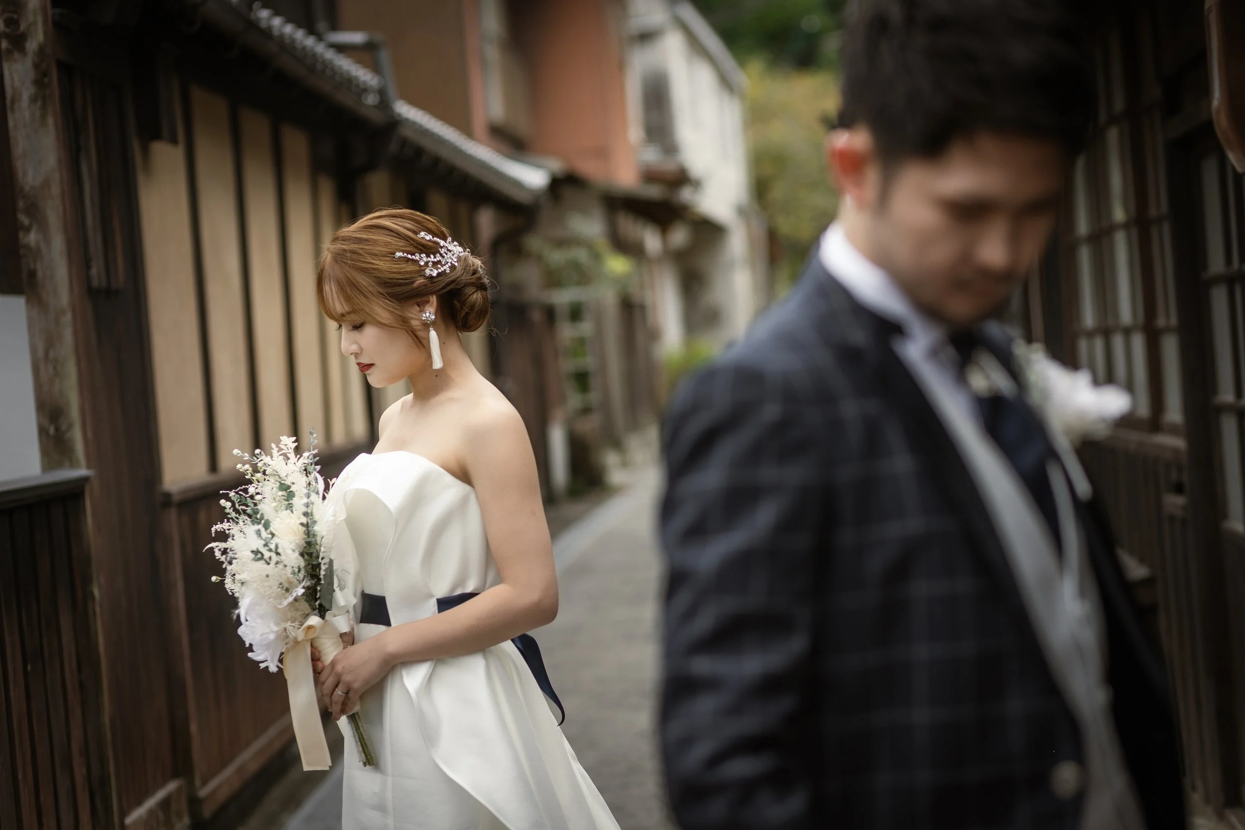 ロケ撮影イメージ | 御手洗町並み保存地区 | 婚礼のドレスを着た女性とタキシードの男性が古い木造の町並みの路地に立っている。彼女は花束を持ち、男性は遠くを見ている。