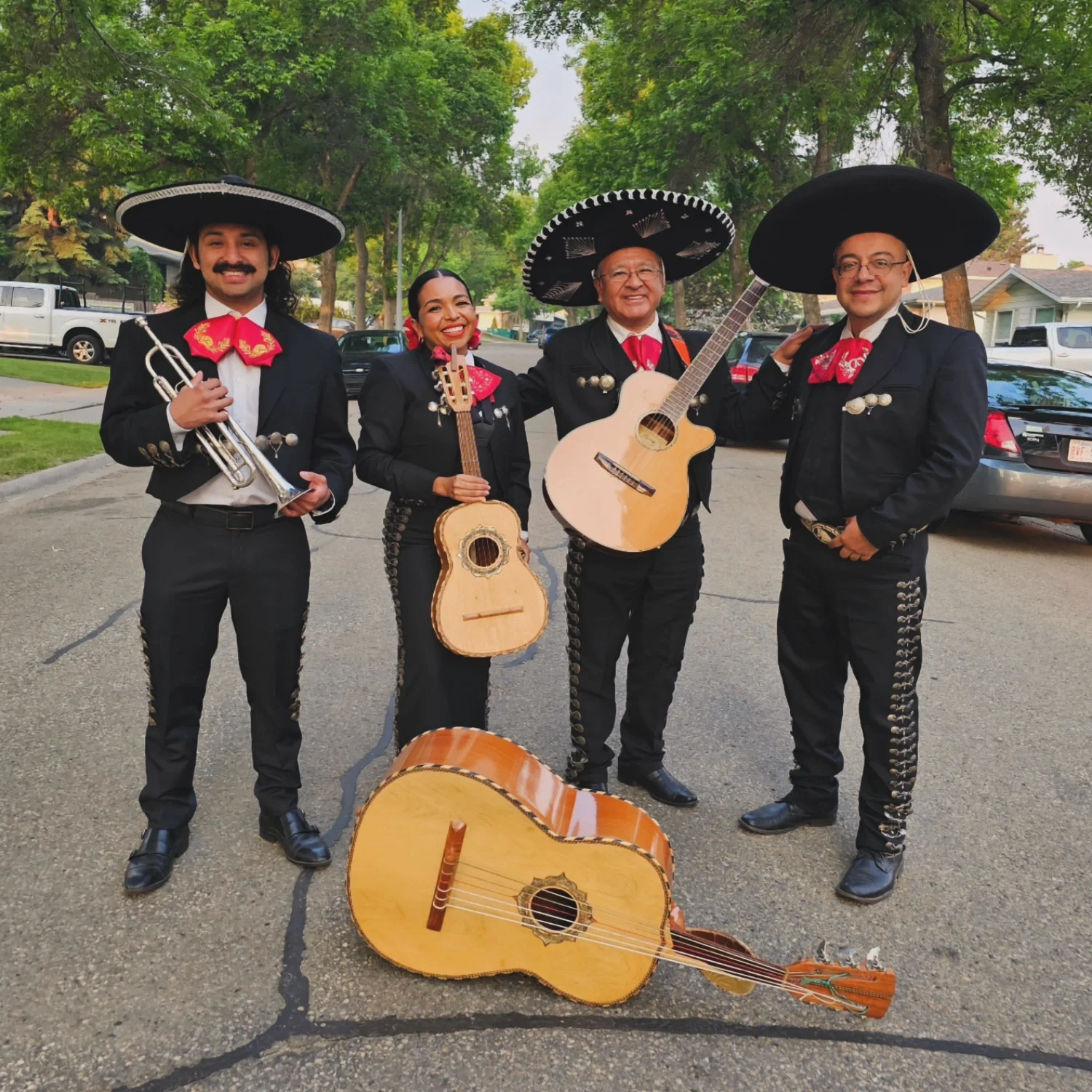 Four musicians dressed in traditional Mexican mariachi outfits, wearing large sombreros, standing on a street with trees and parked cars, smiling. One holds a trumpet, two hold guitars, and one stands next to a guitar that's lying on the ground.