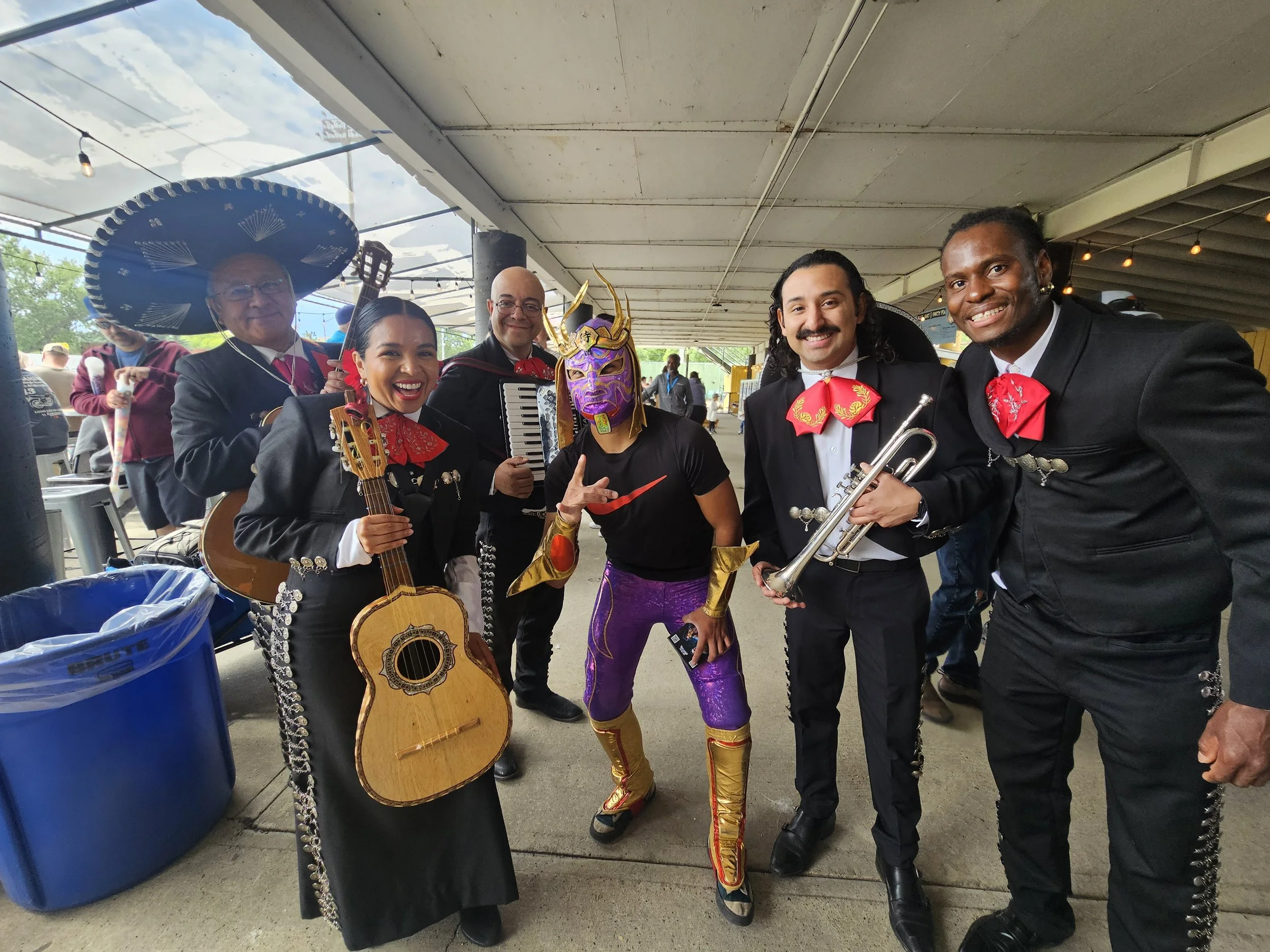Six band members dressed in traditional and costume outfits, holding musical instruments, smiling in an outdoor covered area.