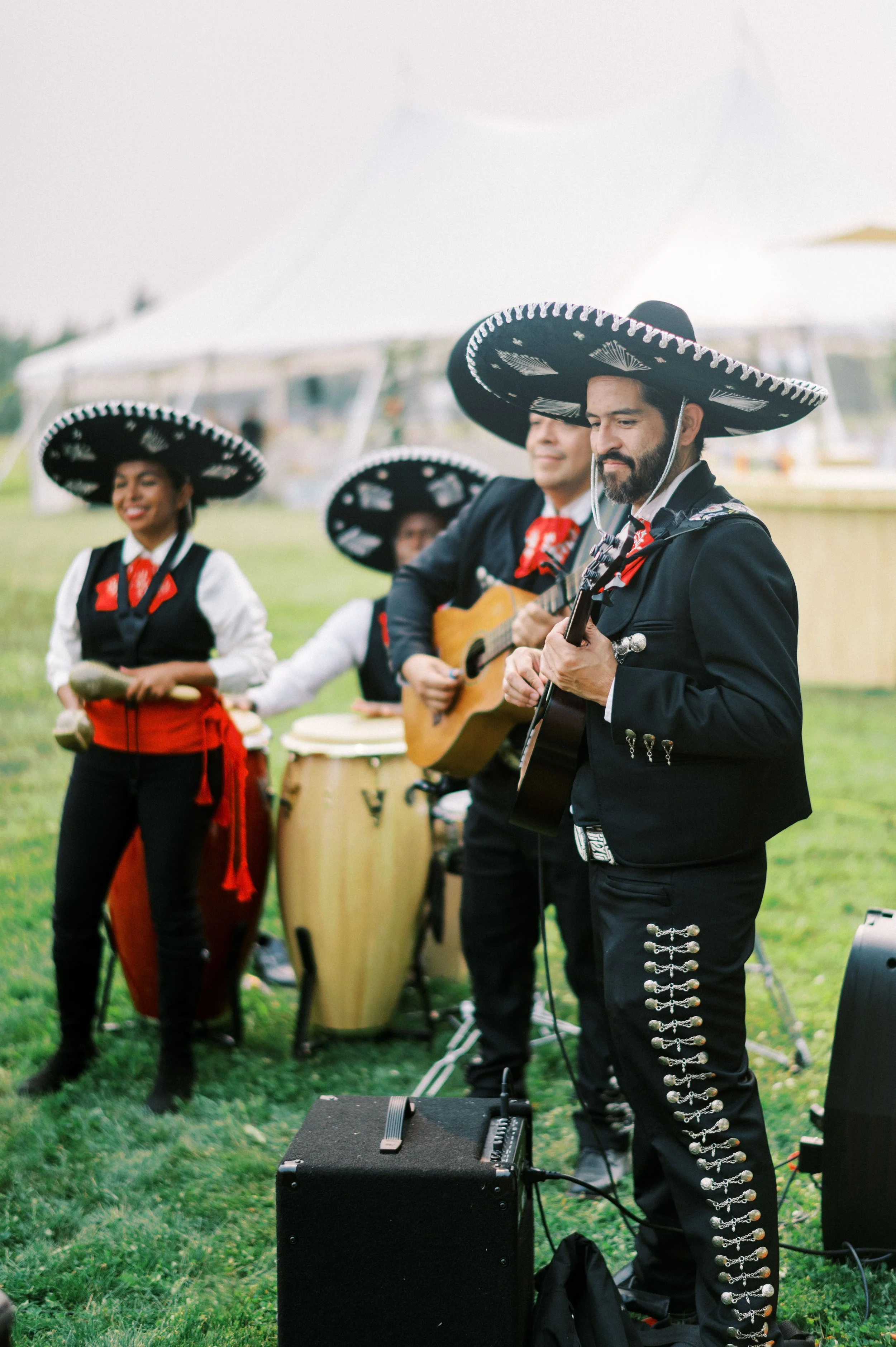 Mariachi musicians performing outdoors under a white tent, wearing traditional black charro suits with silver embroidery and large sombreros, playing guitars, bongos, and maracas.