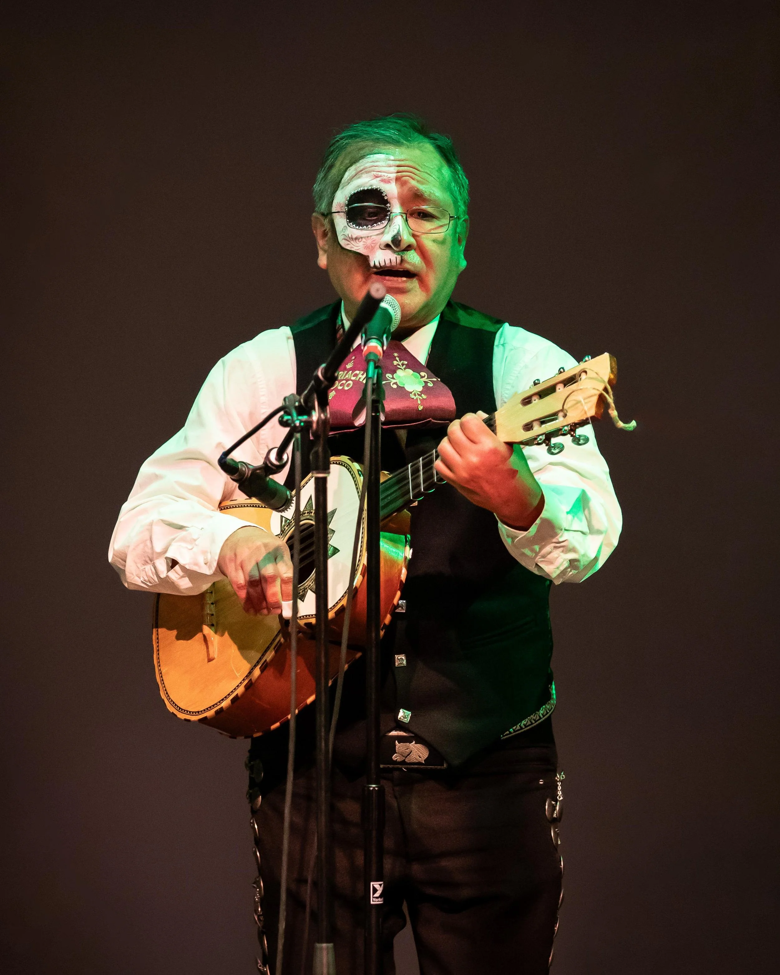 Man dressed in traditional Mexican attire, with half his face painted as a sugar skull, singing into a microphone while playing an acoustic guitar on stage.