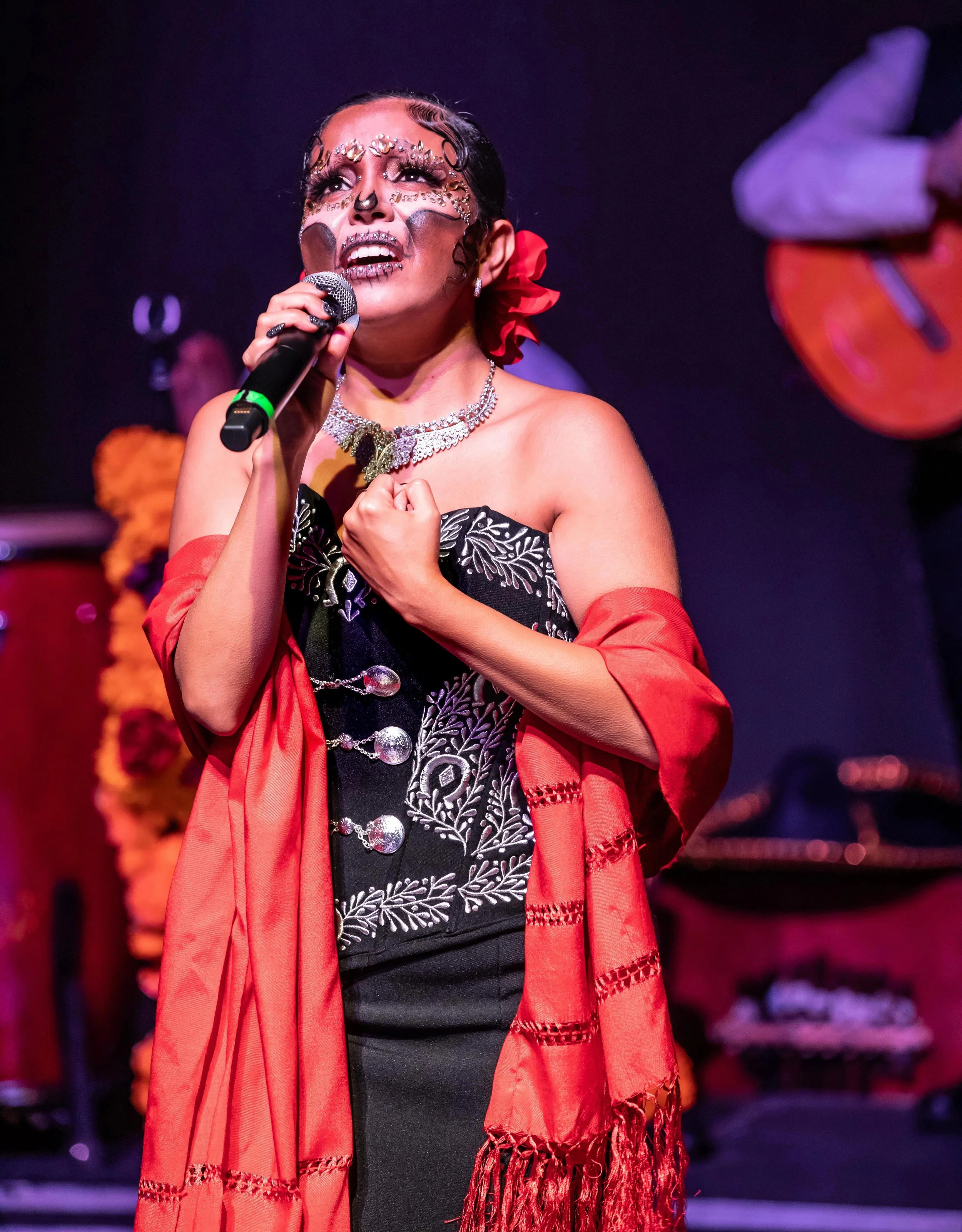 A woman singing passionately on stage with dramatic makeup, jewelry, and a red shawl, holding a microphone