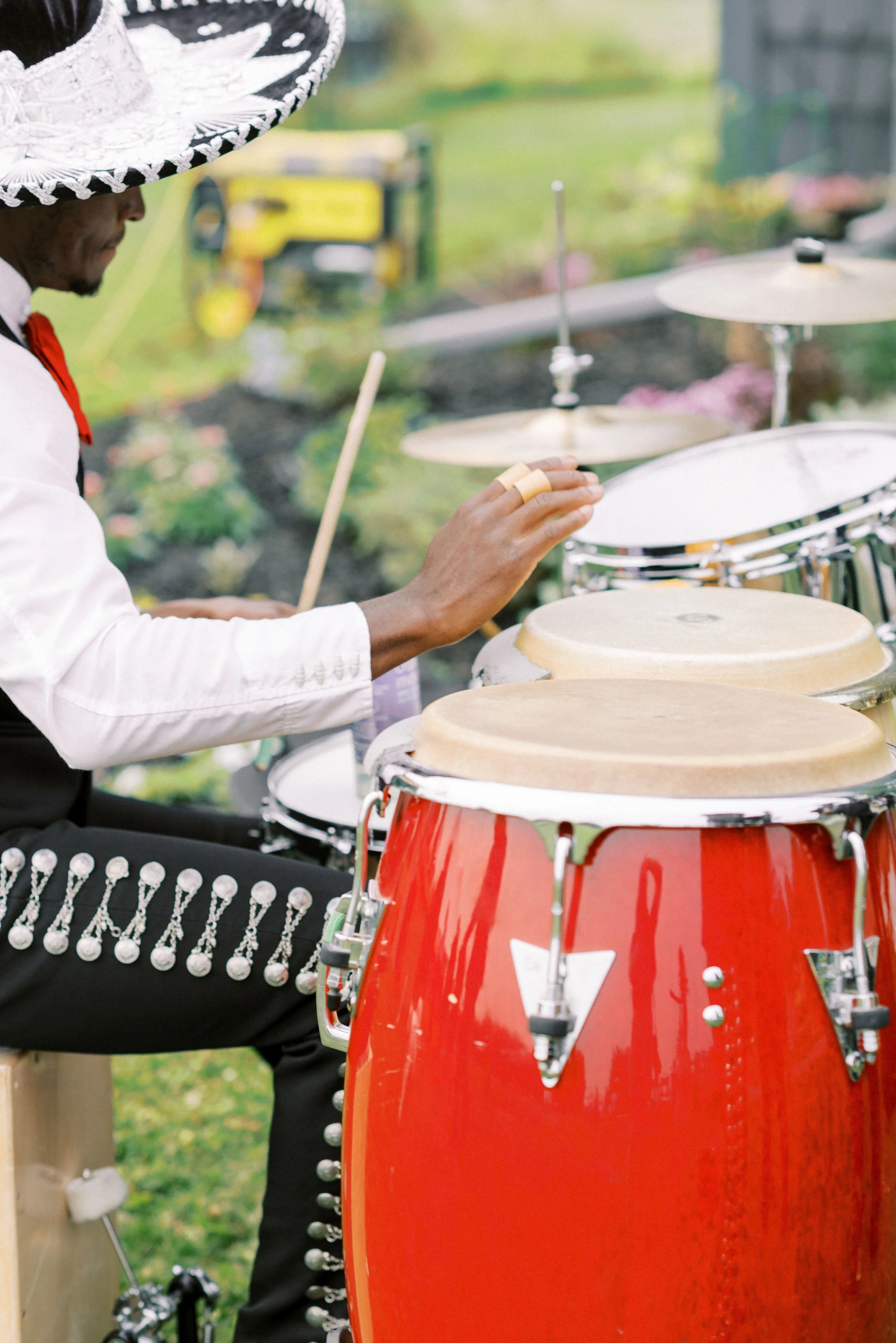 A person playing percussion instruments outdoors, wearing a large hat, white shirt, and black pants with silver embellishments.