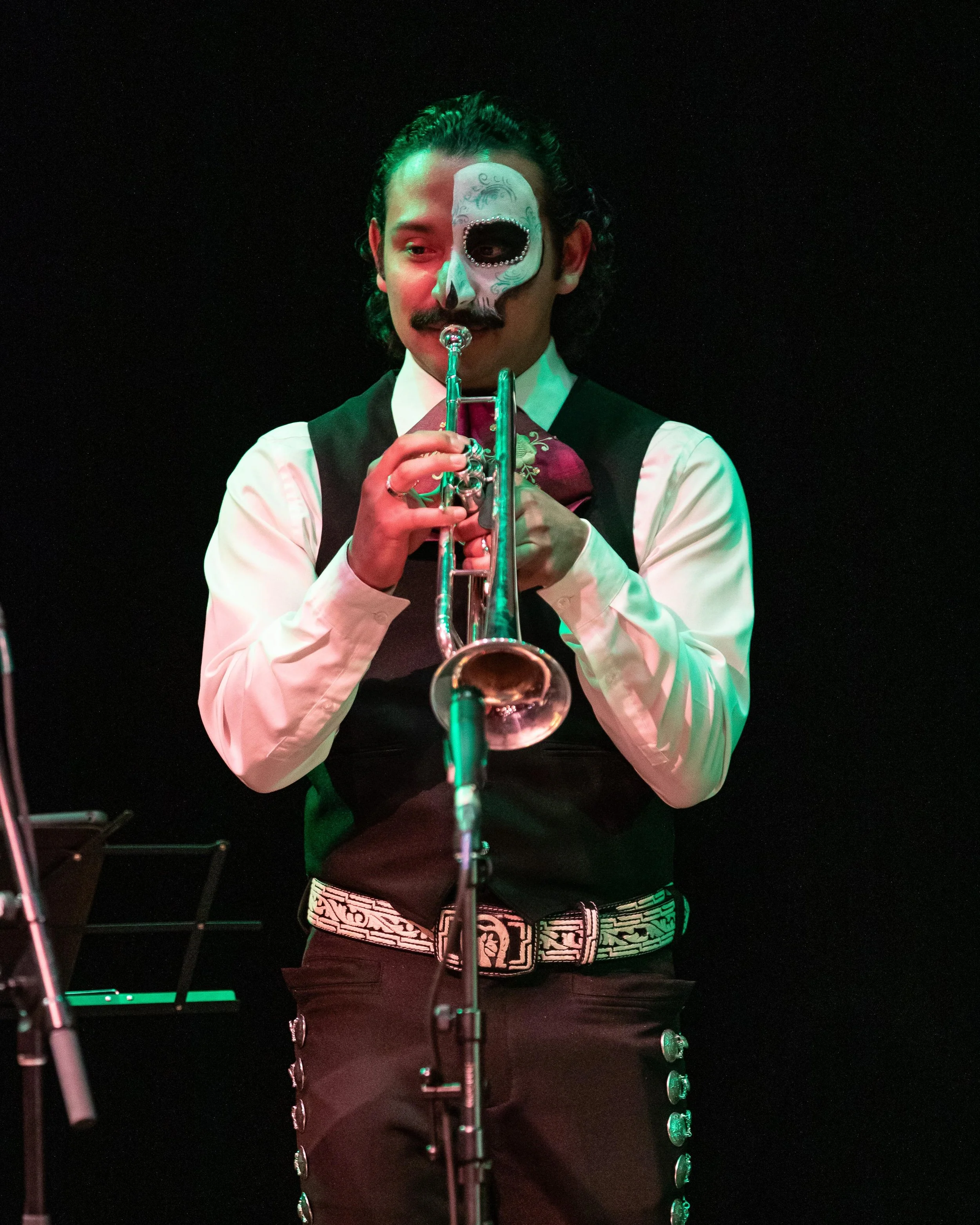 A man in costume with half his face painted as a skeleton, playing a trumpet on stage, wearing a white shirt, black vest, and a patterned belt.