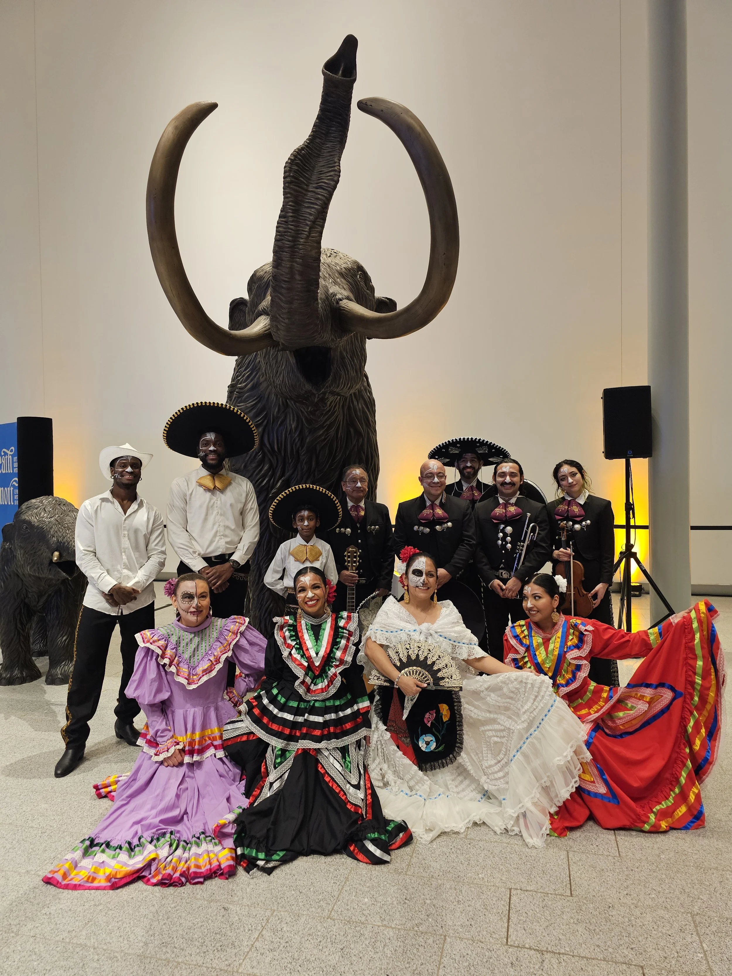 A group of dancers and musicians dressed in traditional Mexican attire standing in front of a large elephant sculpture with tusks.