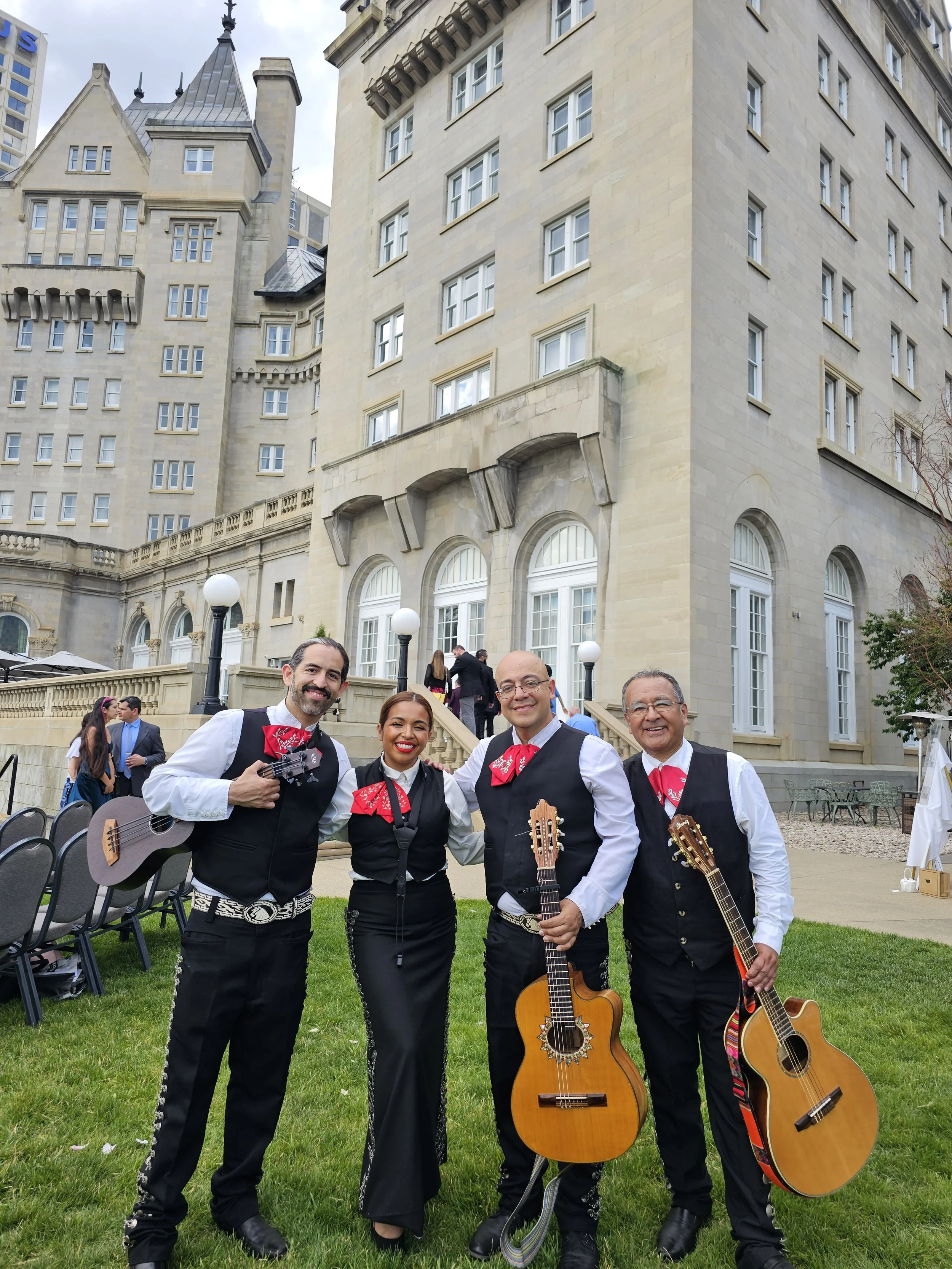 Four mariachi musicians standing outdoors on green grass with a historic building in the background. Three men and one woman are holding guitars, wearing traditional mariachi attire with red bow ties and black vests.