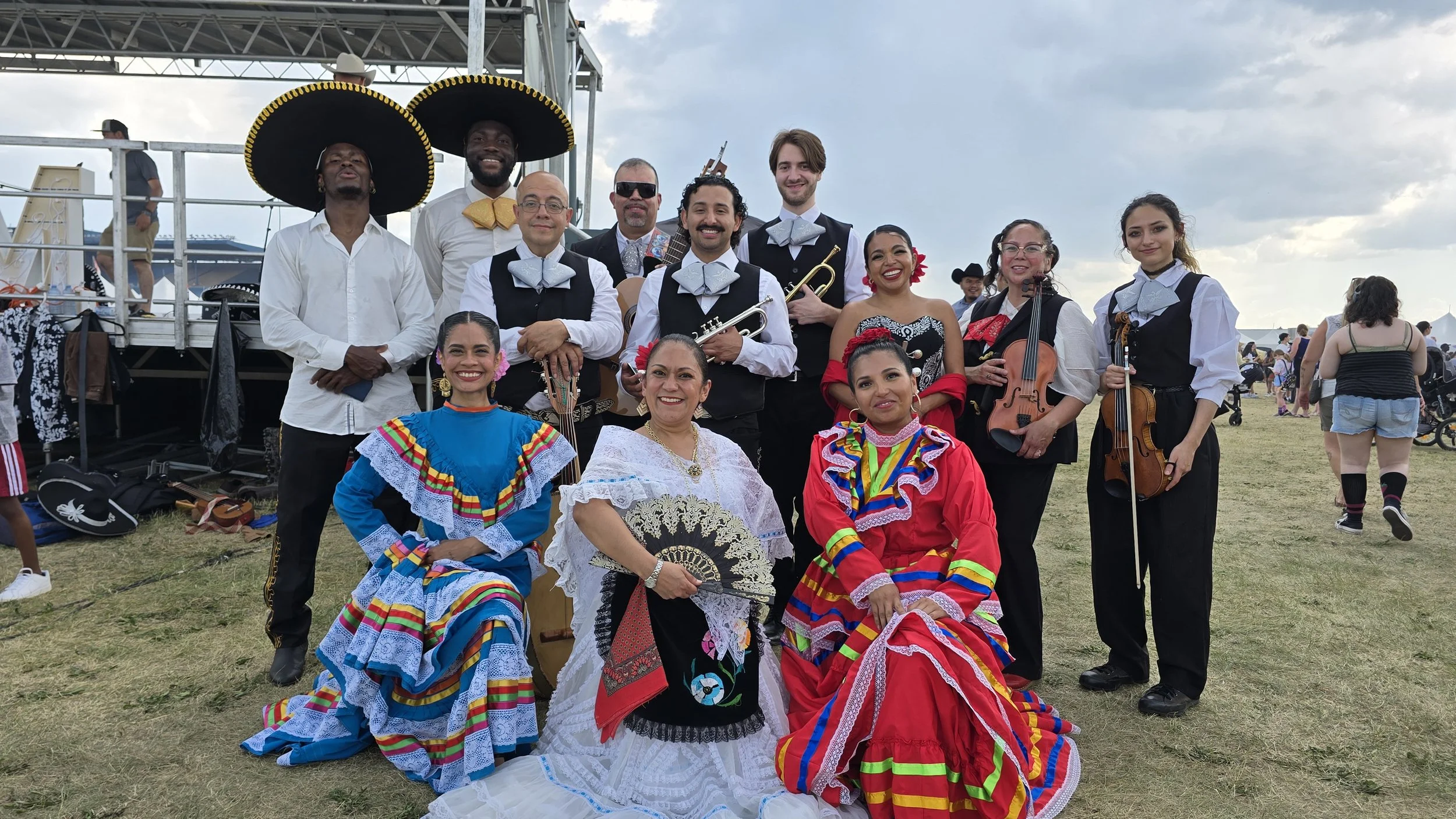Group of musicians and dancers in traditional Mexican attire, some holding instruments like violins, a trumpet, and a guitar, outdoors at a cultural festival.