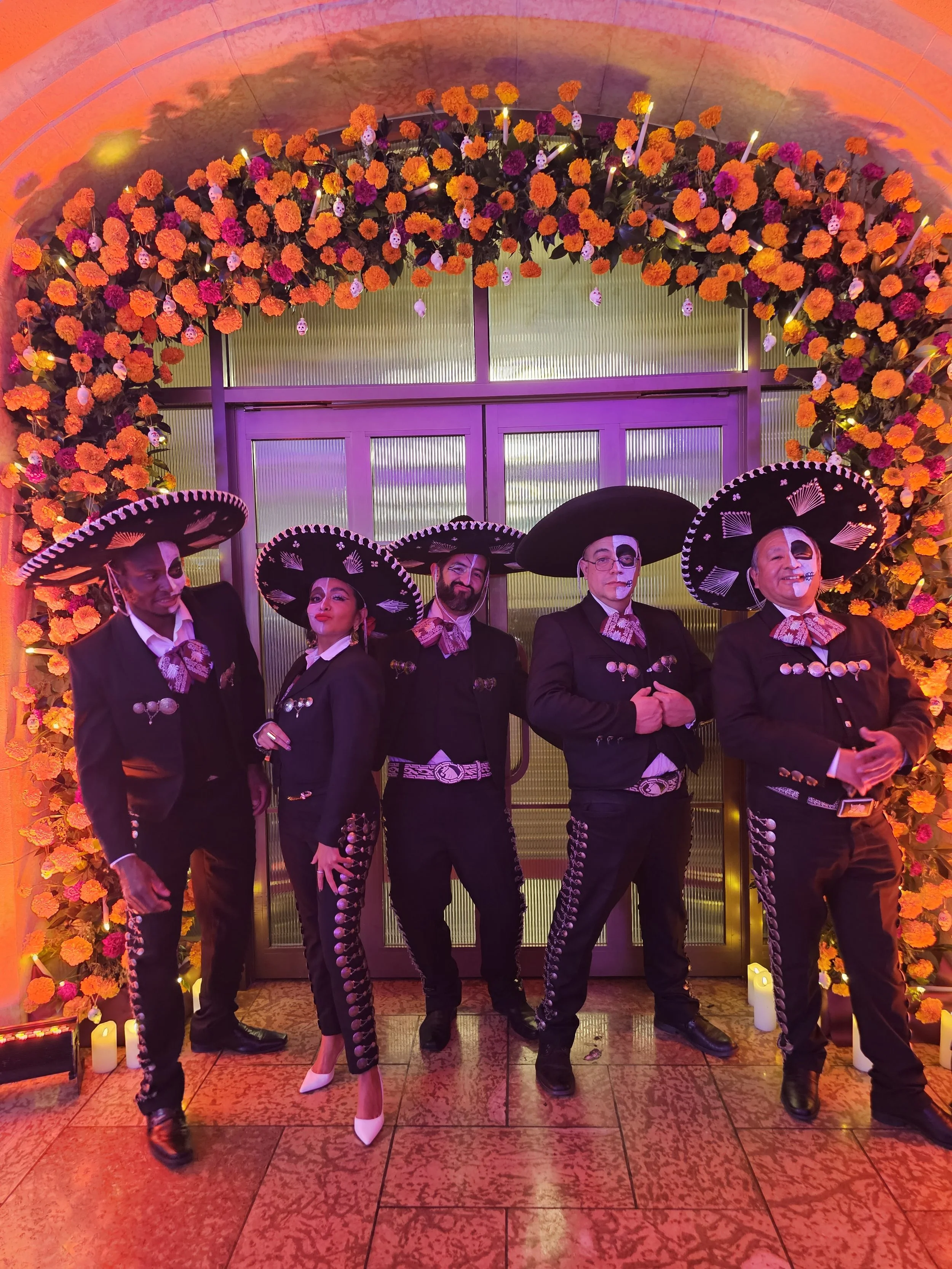 Group of five people dressed as Mexican revolutionary icons, wearing sombreros and traditional costumes, standing under a floral arch with marigolds, lit with purple and yellow lighting.