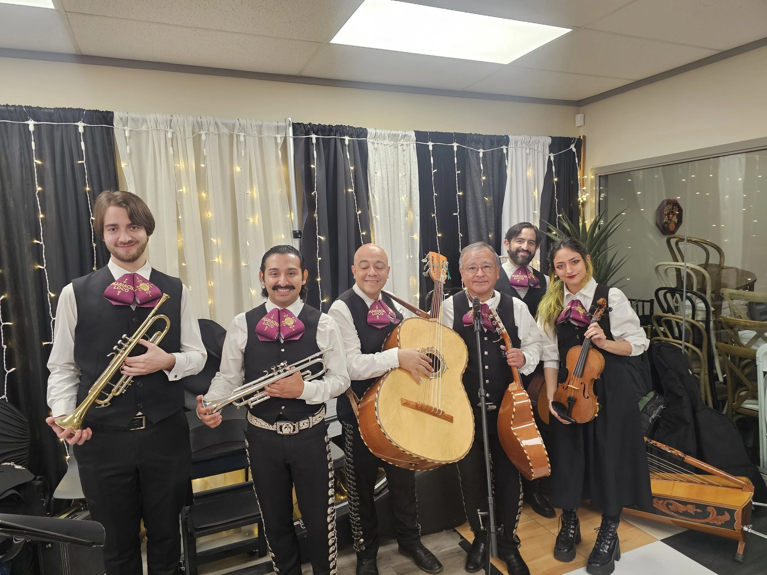 Group of six musicians in formal attire holding various string and wind instruments, standing inside a decorated room with string lights and curtains.