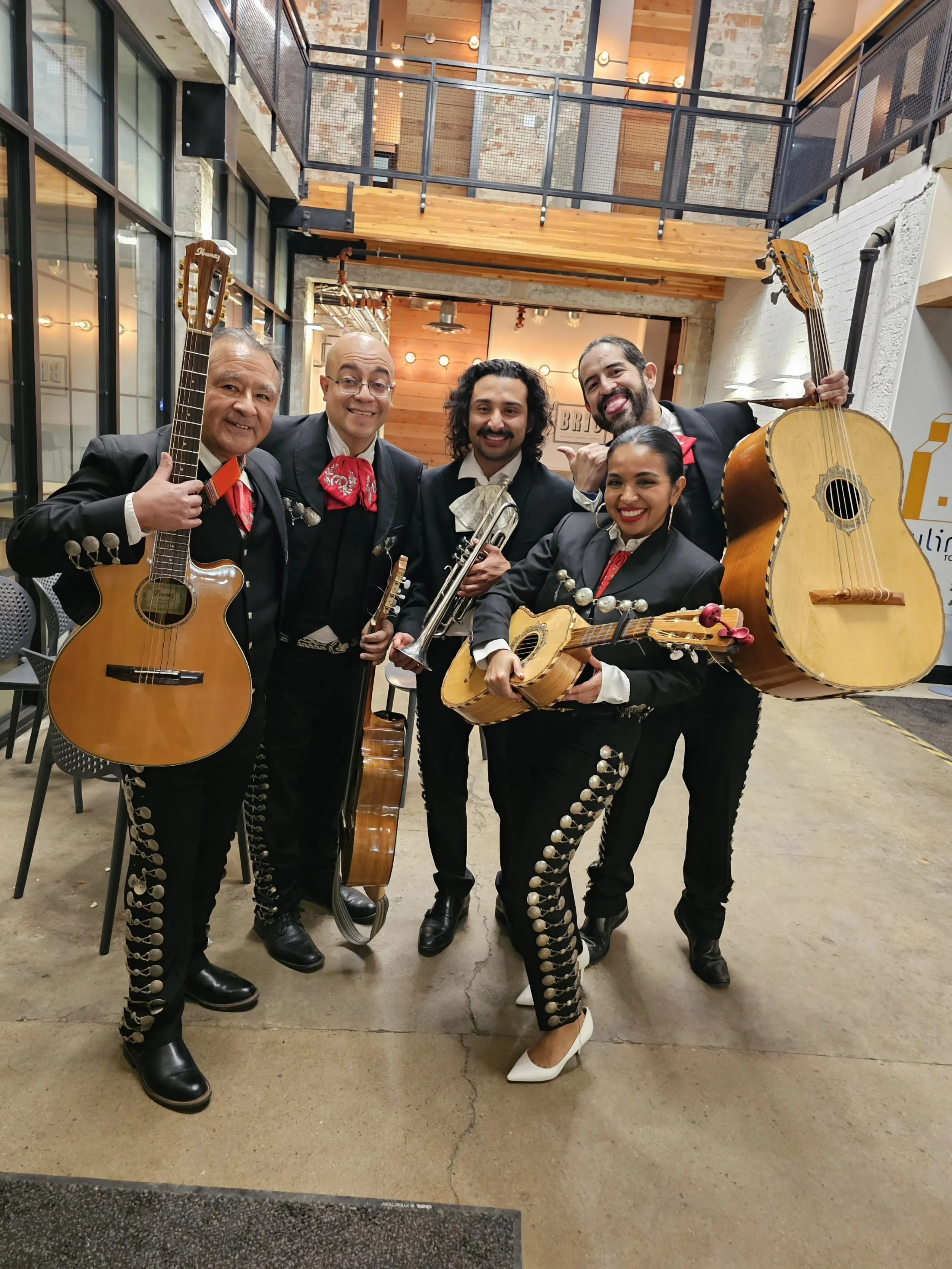 A group of six mariachi musicians posing indoors with their instruments, wearing traditional black outfits with silver accents and red ties.