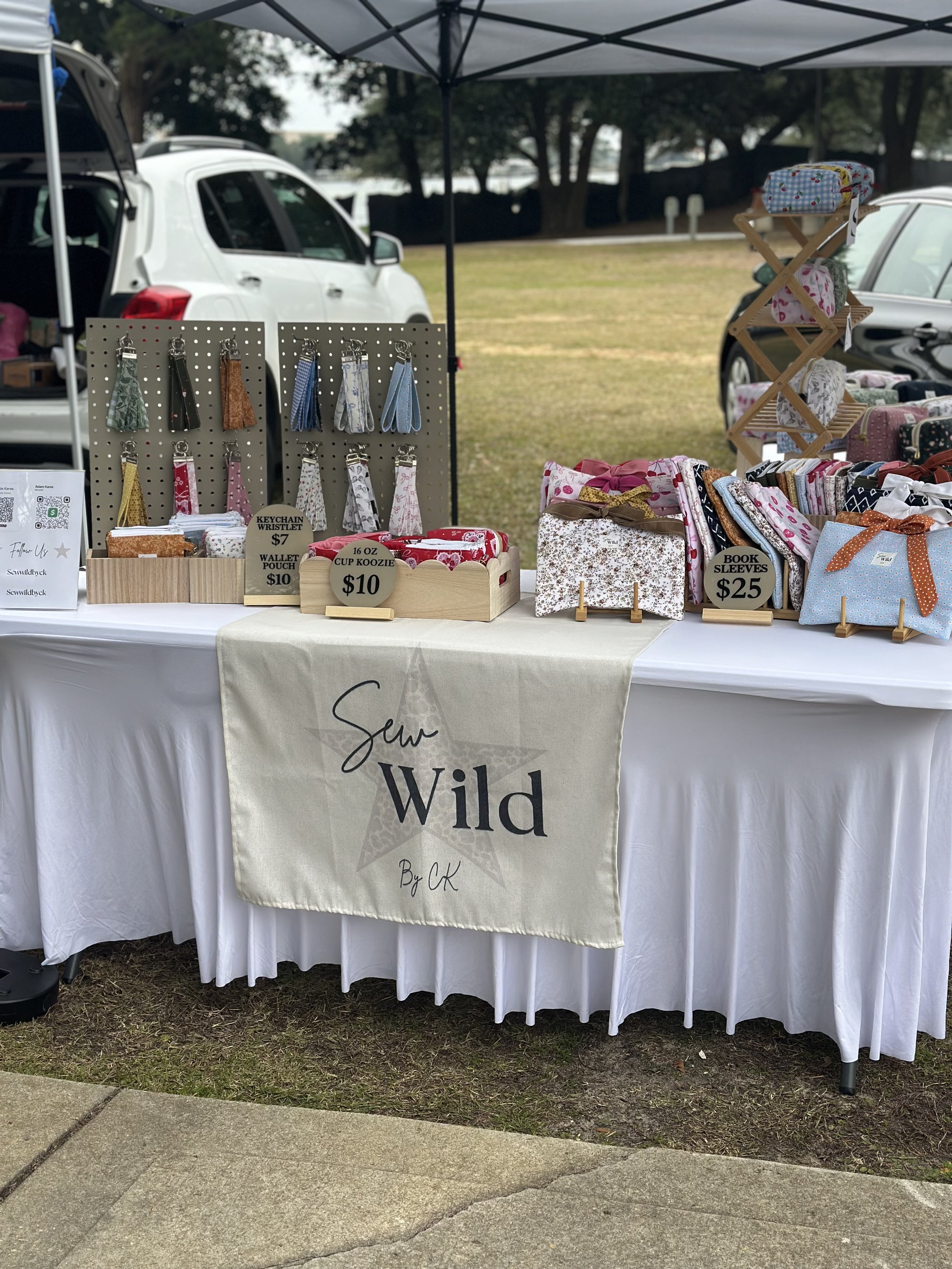 A market stall called 'Sew Wild By CK' displays fabric hair ties, keychain wristlets, wallet pouches, cup koozies, and book sleeves, with cars and trees in the background.