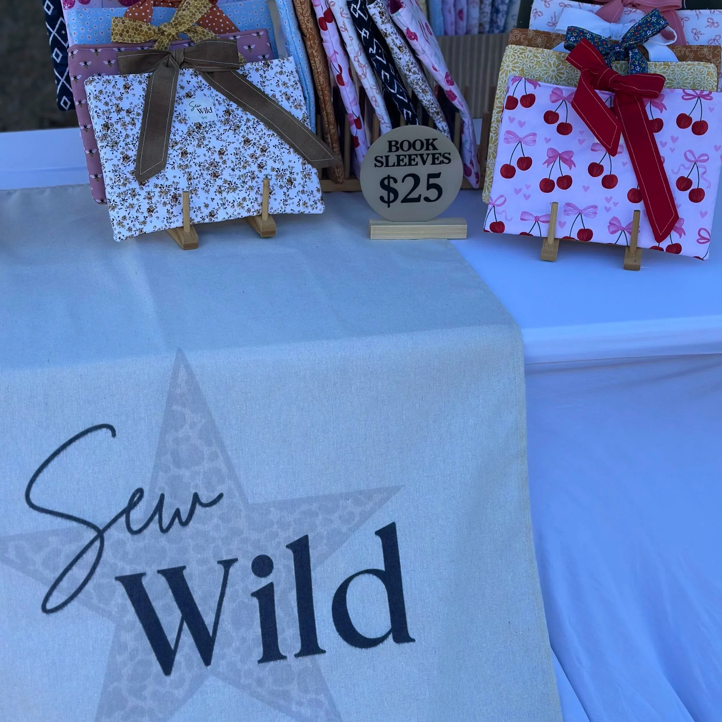 Display of fabric book sleeves with floral and cherry patterns, priced at $25 each, on a table with a cloth that has a logo reading 'Sun Wild' and a stylized star. The sewing supplies and patterns are arranged behind the sleeves.