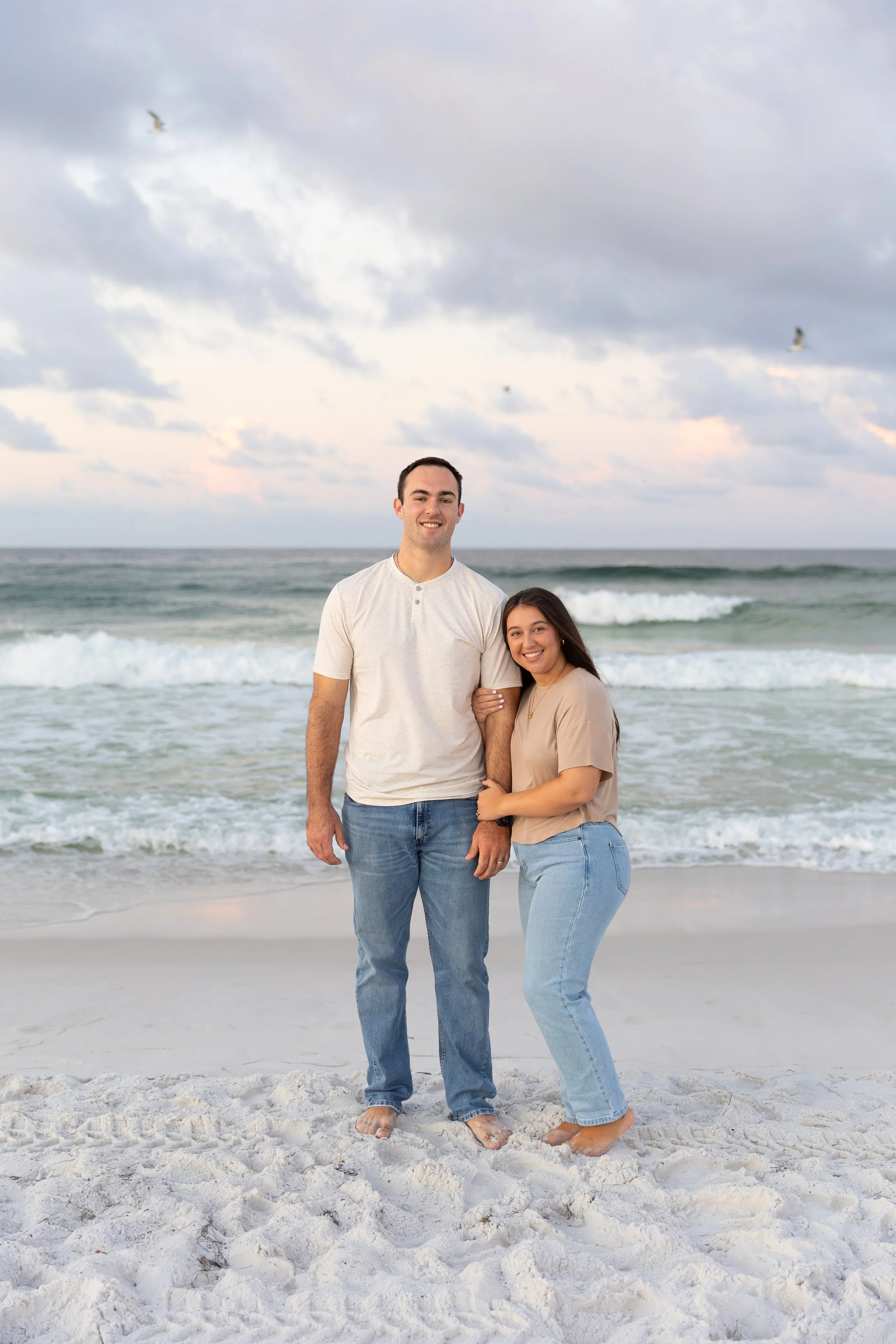 A young couple standing on a sandy beach with ocean waves and a cloudy sky behind them, smiling at the camera.