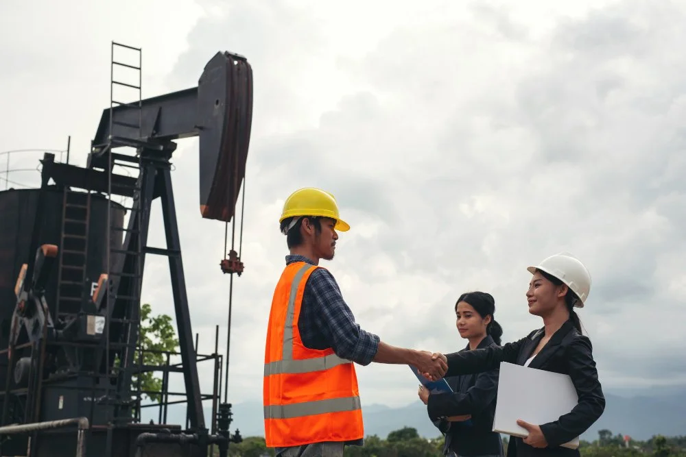 Three engineers or construction workers wearing safety helmets, two women and one man, are outdoors at an oil or gas drilling site. They are shaking hands, and one woman is holding a clipboard and a laptop.