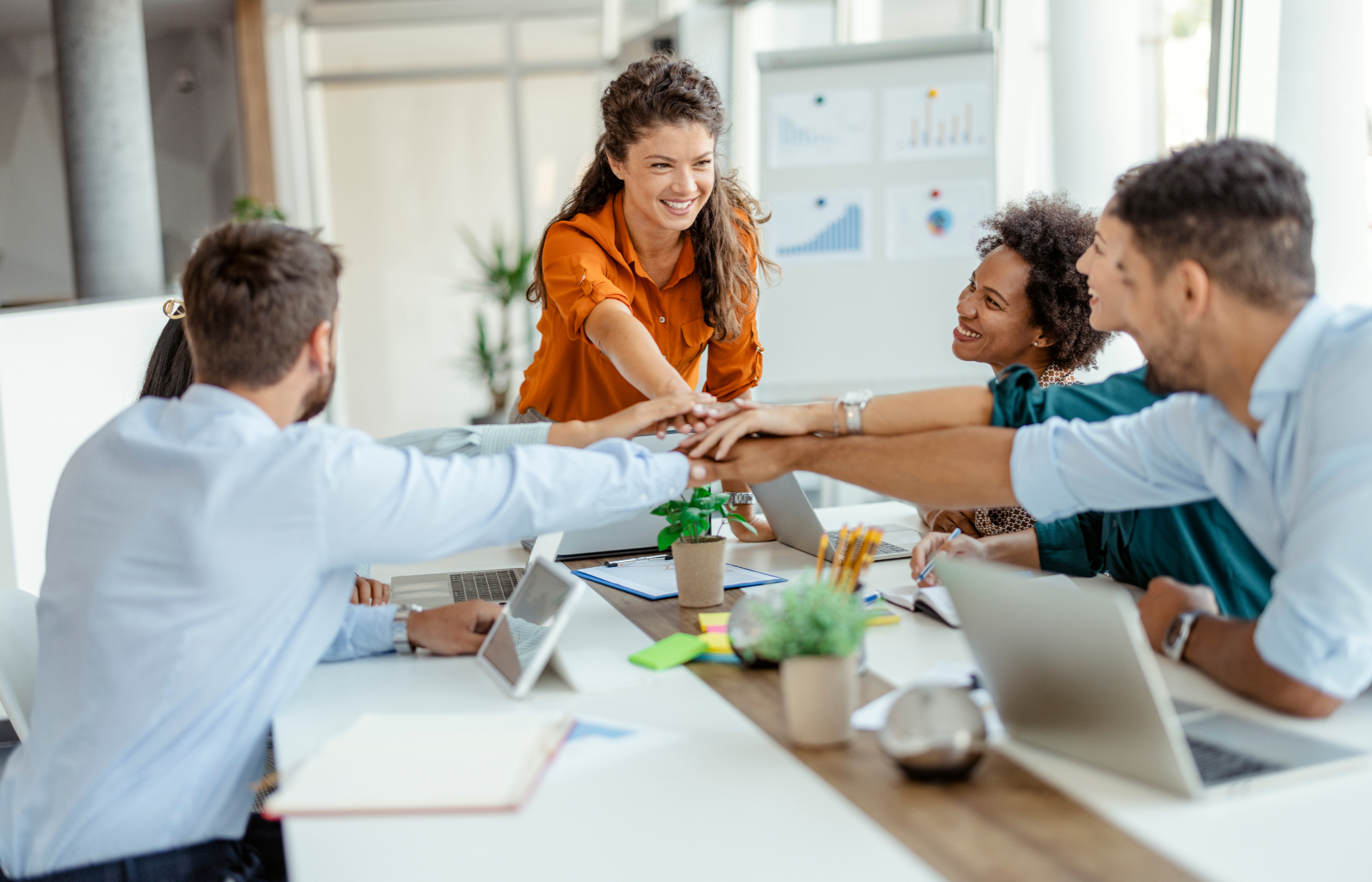 A group of five diverse colleagues in a business meeting, smiling and stacking their hands together in a gesture of teamwork and unity inside a bright conference room.