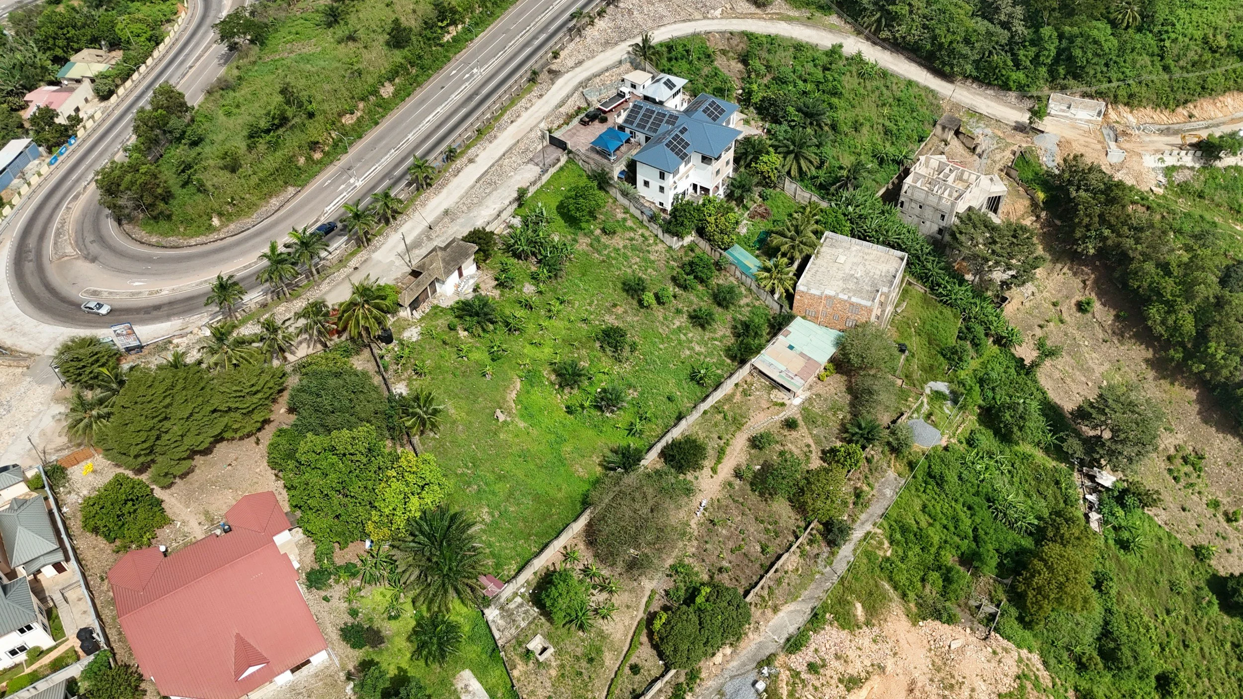 Aerial view of a residential area with houses, a curved road with cars, green vegetation, and open plots of land.