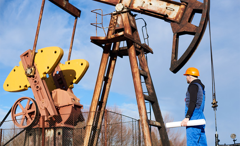 A worker wearing a blue uniform and orange safety helmet holding a blueprint, standing next to large rusted oil pumpjack equipment outdoors against a blue sky.