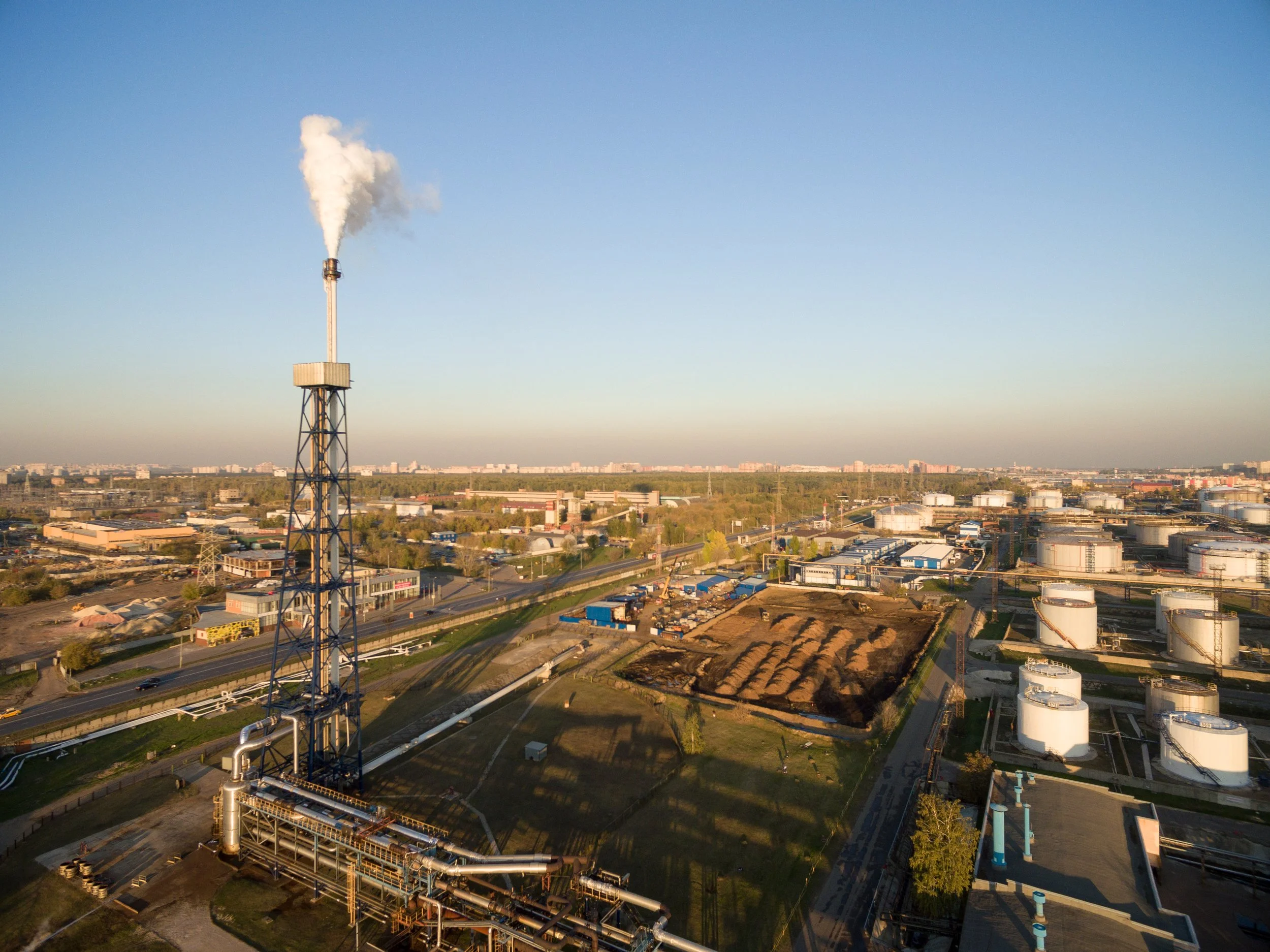 An industrial plant with a tall smokestack emitting white smoke, surrounded by storage tanks and buildings, under a clear blue sky.